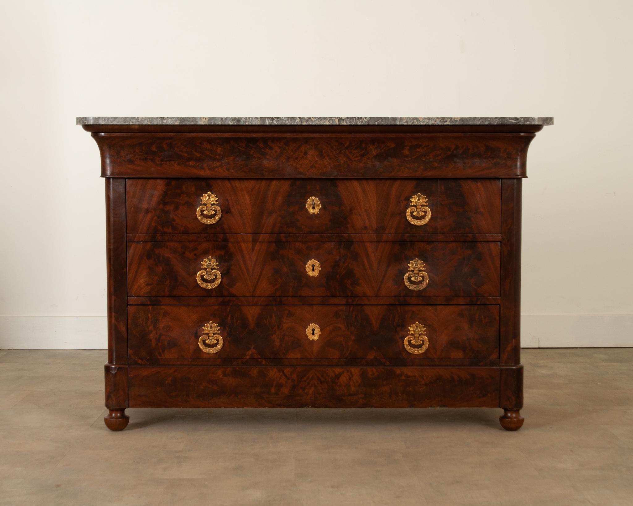 This Louis Philippe style commode is crafted from stunning burl mahogany. The charcoal and white St Anne Gris marble top is original to the piece and has rounded front corners. The apron houses a hidden drawer atop three of the strikingly