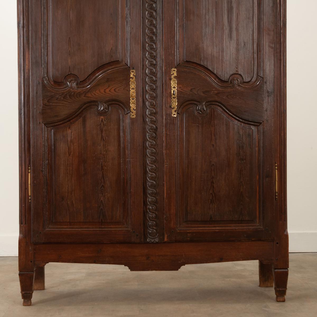 This French armoire from circa 1890 is full of show-stopping carvings. A wonderfully rounded cornice sits above a pair of carved paneled doors that are bisected by an eye-catching, twisting motif.  Interesting brass escutcheons depict hearts and