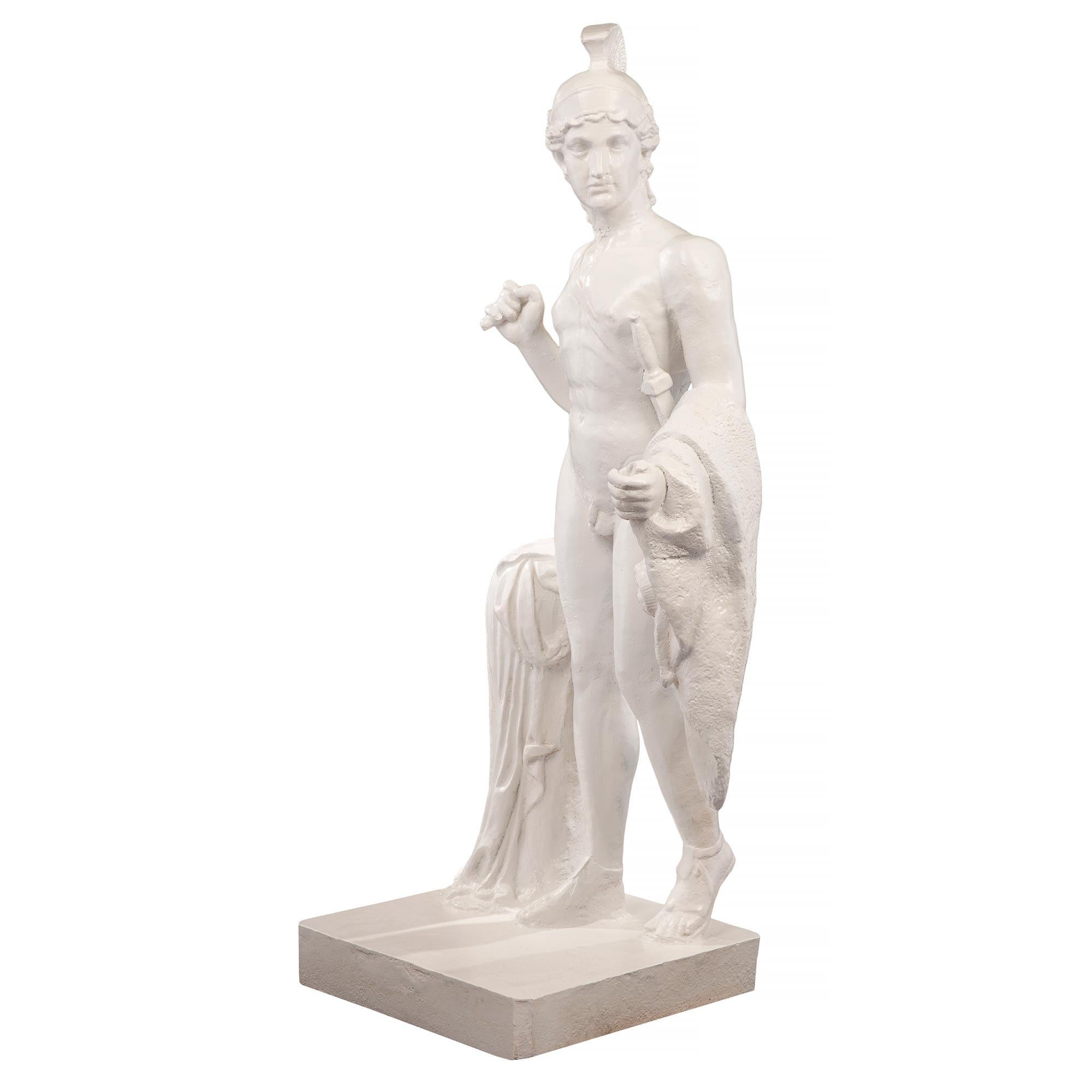 Statue en fonte du XIXe siècle représentant un jeune centurion. La statue est surélevée par une base carrée où il se tient à côté d'une souche d'arbre. Le beau personnage se tient debout, sa cape rehaussée d'un trophée à tête de bélier drapée sur
