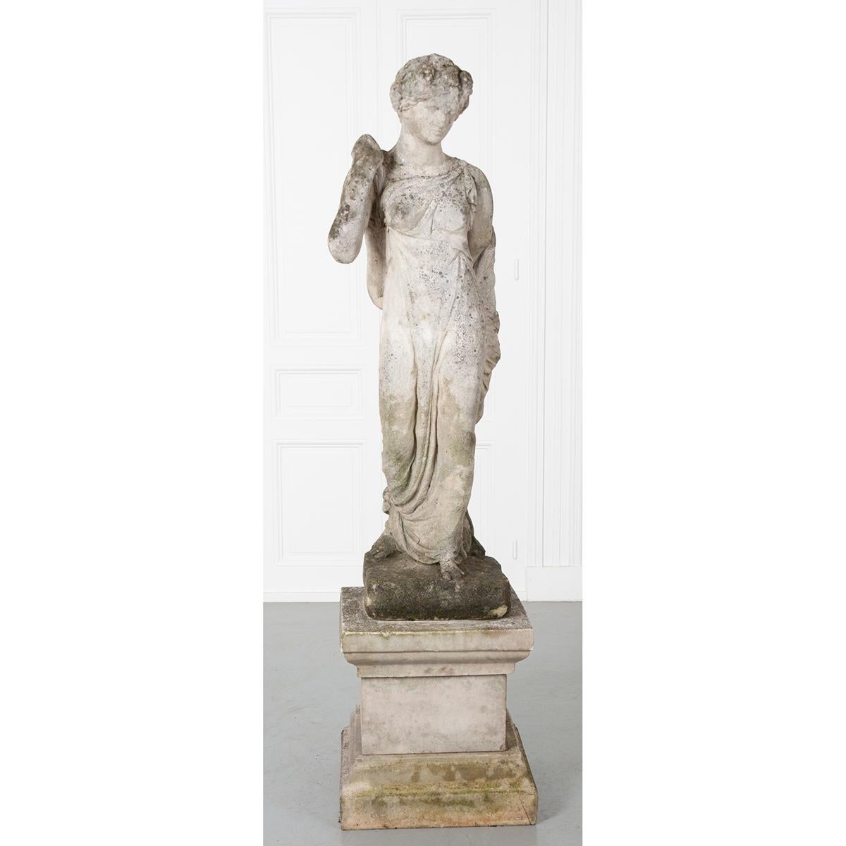 This patinated cast stone statue rests atop a pedestal that would work beautifully indoors or outdoors. It depicts a robed female with one hand tucked into her robe with the other hand over her shoulder. Peeking out from under her robe you can see