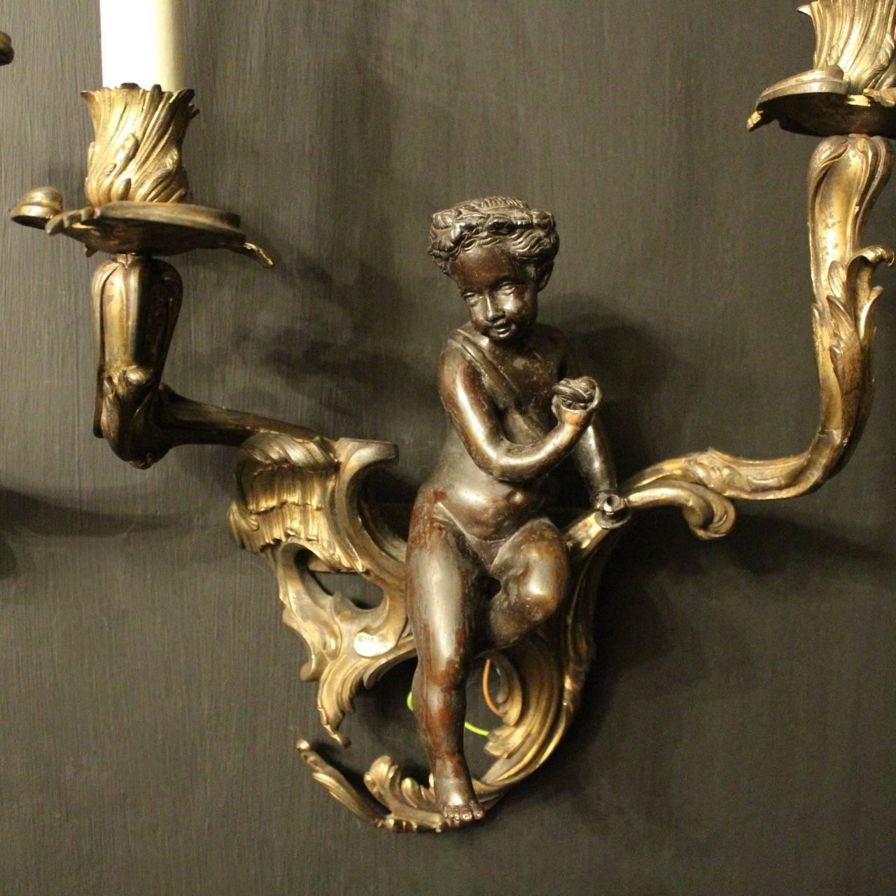 A French pair of gilded bronze twin arm antique wall sconces, the ornate leaf scrolling arms with leaf bobeches drip pans and bulbous candle sconces, issuing from a decorative leaf pierced scrolling backplate with lovely central cherubs, great
