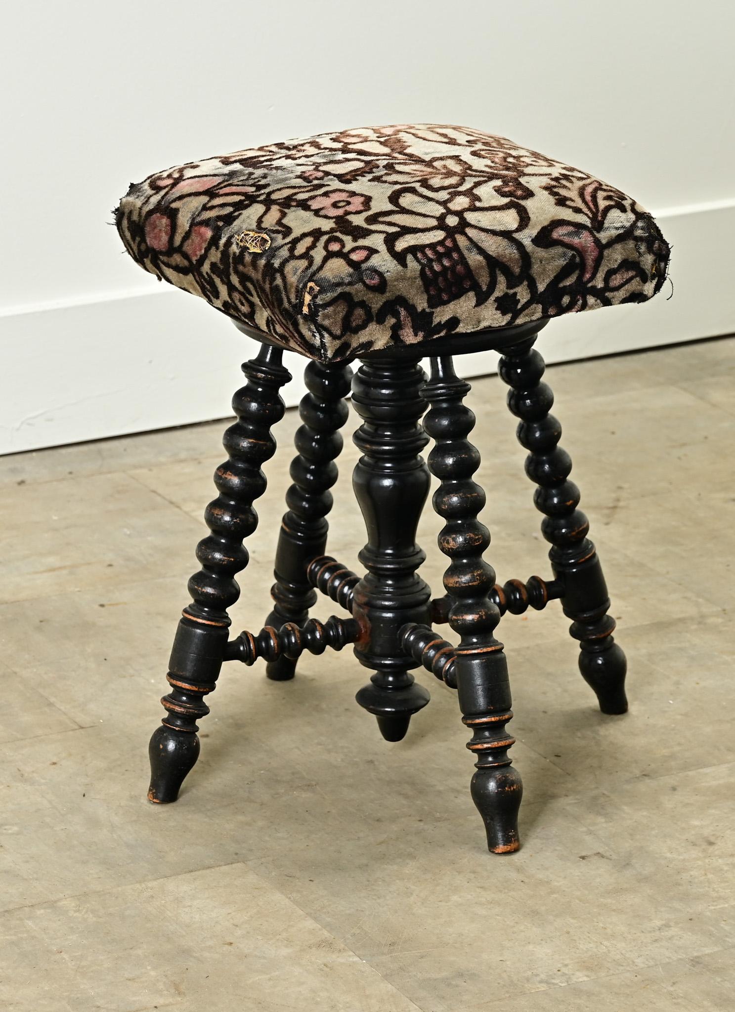 A charming upholstered and ebonized piano stool made in France. The top cushion is upholstered in a worn floral-print velvet that compliments the ebonized wooden frame. The center of the stool has a hidden steel screw used to raise and lower the