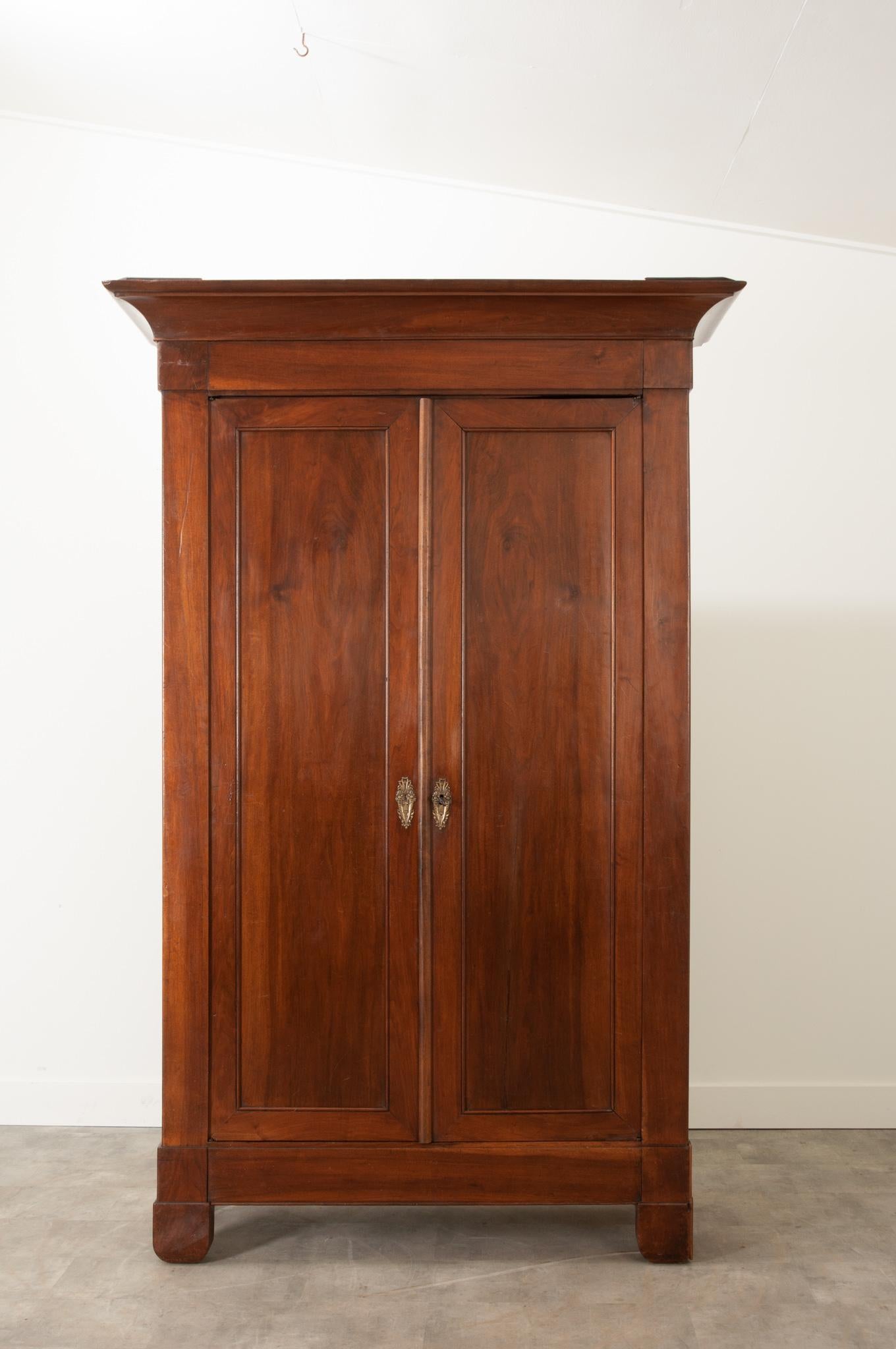 This handsome Empire style walnut armoire features a carved cornice over a pair of paneled doors mounted with stylized brass floral motif escutcheons. Recently polished with French wax paste to revive the rich tone and patina of the wood. A single