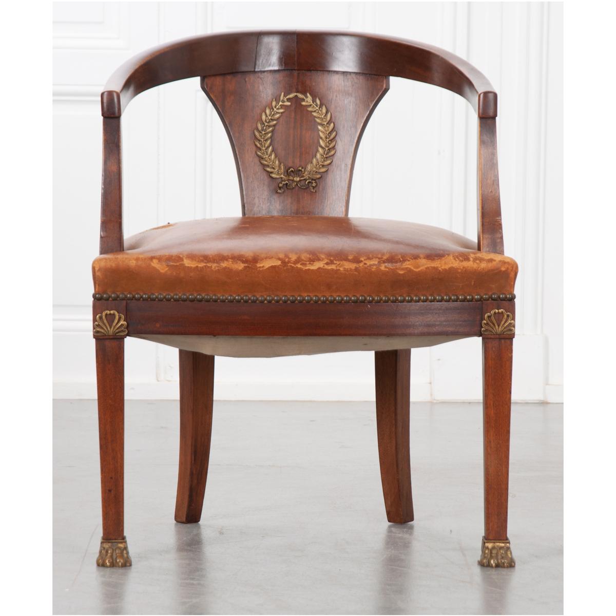 A handsome Empire barrel back chair. This mahogany chair has a brown/tan antique leather upholstered cushion trimmed with a nailhead border. The concave back has a shaped backrest with a large ormolu laurel wreath at its center. The back extends