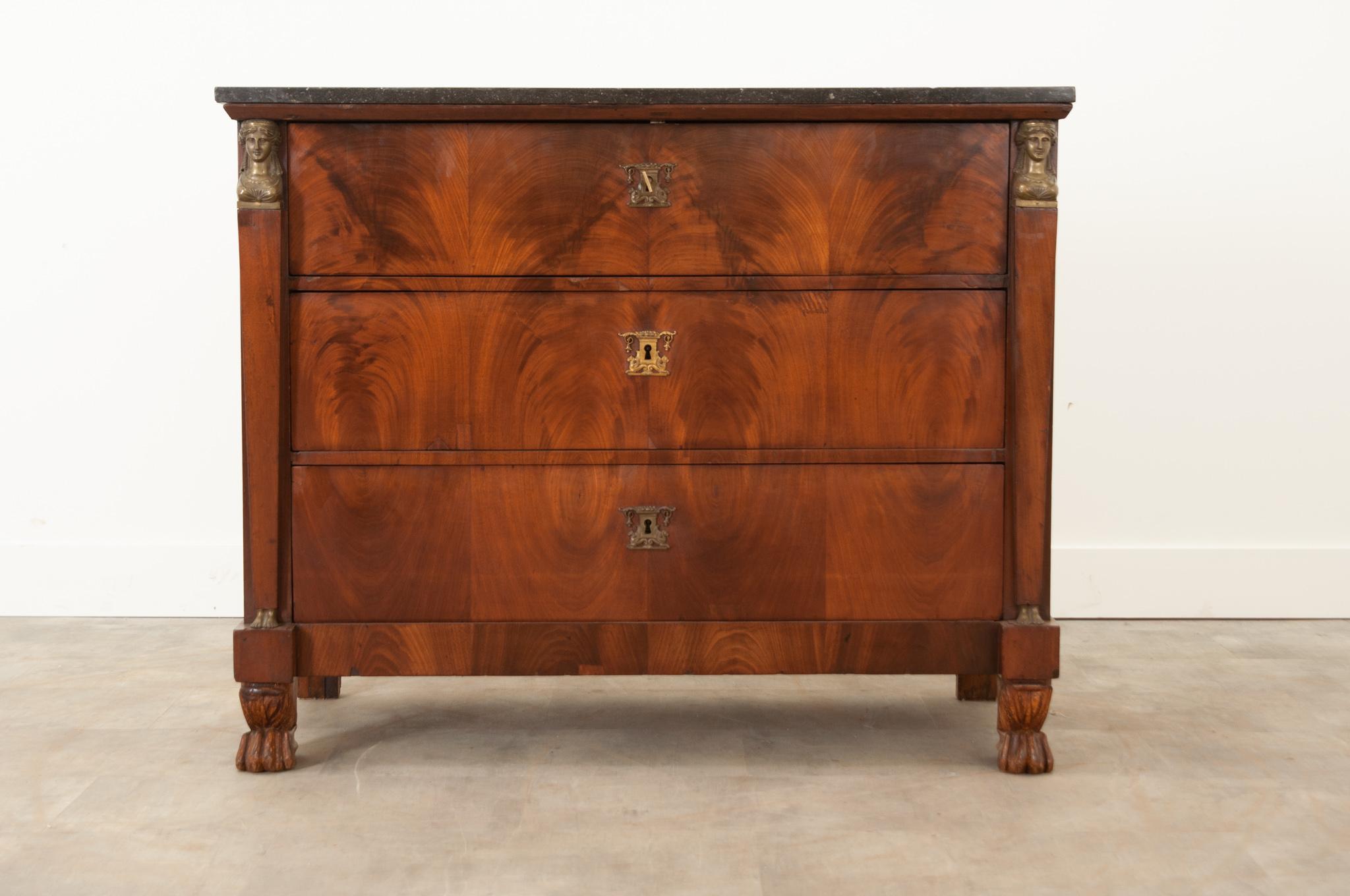 This French 19th century commode features fantastic Empire style elements. Topped with a removable piece of black fossil marble that features a few worn nicks around its outer edge. All three bookmatched mahogany drawers feature matching cast brass