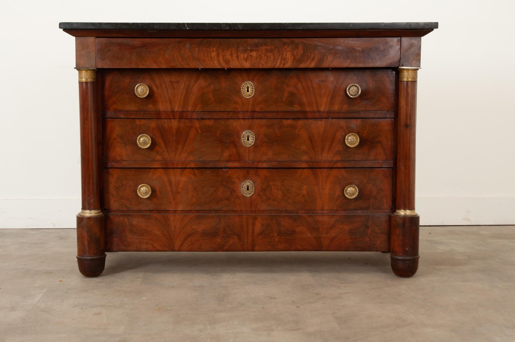 A classic French Empire commode topped with its original black marble. This commode is finished in warm bookmatched burl mahogany. There are four drawers total. At the top is a single drawer opened with a hidden under-drawer pull. Below are three