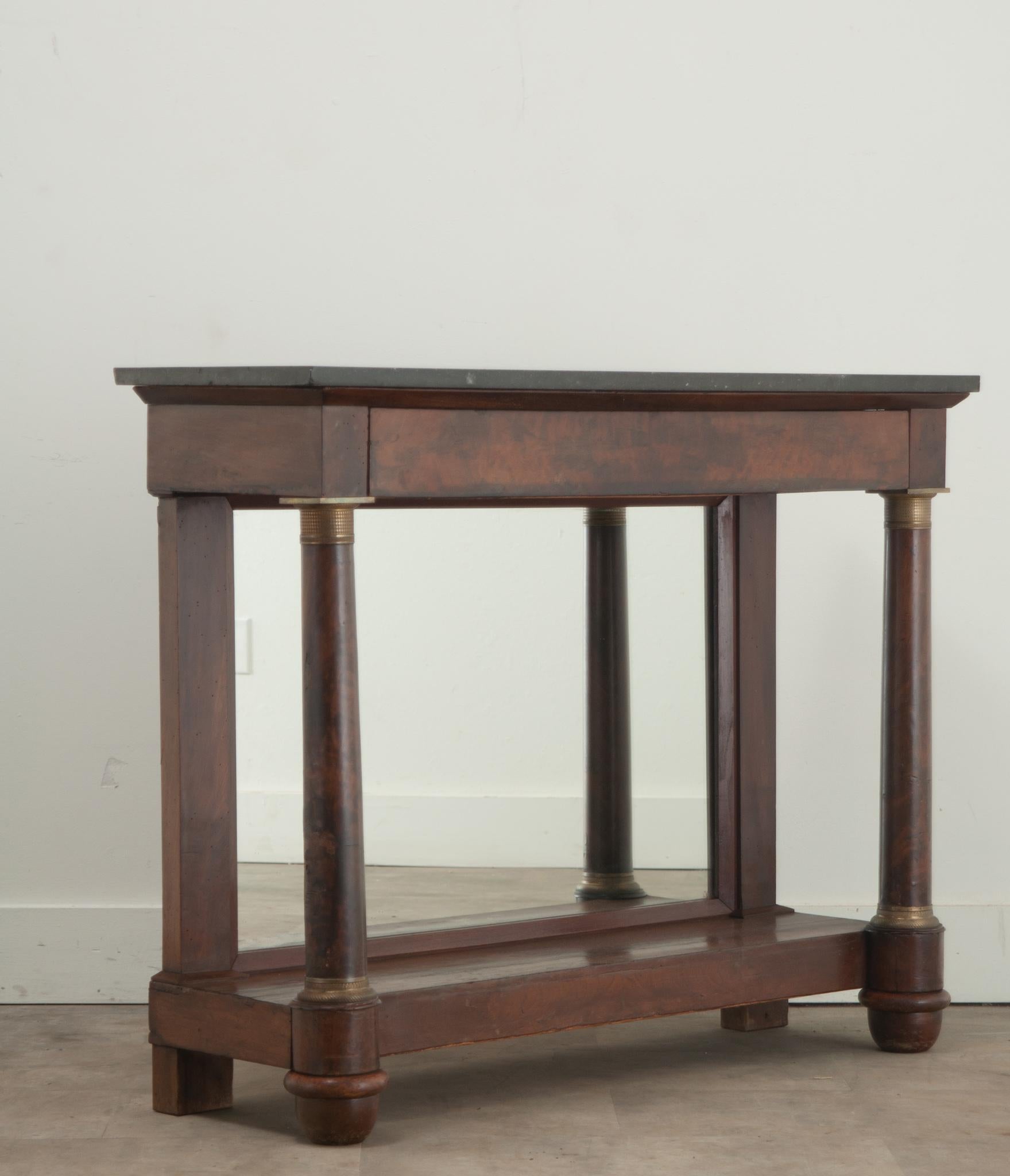 A classic French Empire mahogany console from the 19th Century. The original black fossil marble top is filled with shells and other petrified sea life. The simple apron contains one drawer and is supported by turned column front legs accented with