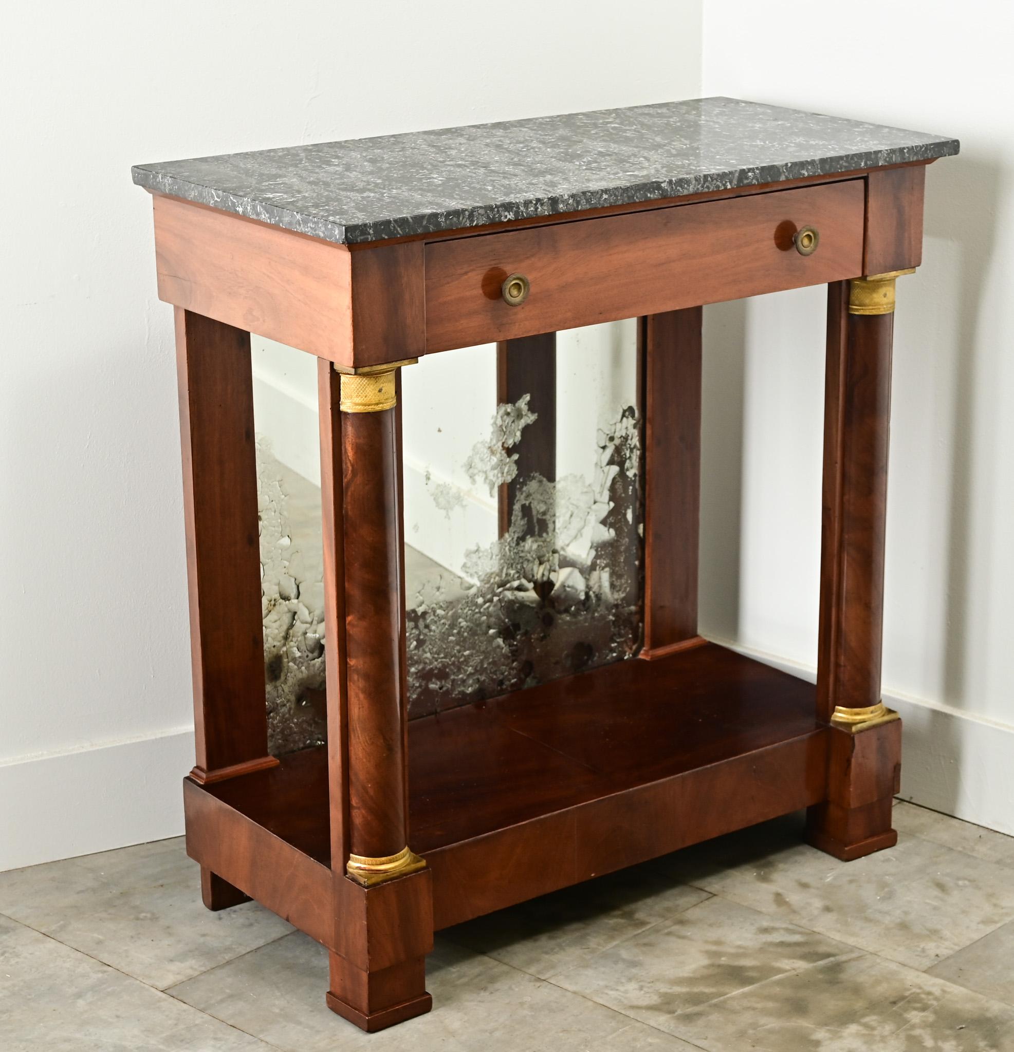 A petite French mahogany Empire console with its original marble top. A single drawer is housed in the apron over a pair of column form front legs with neoclassical style brass capitals at the top and base. Behind the console is the original mirror