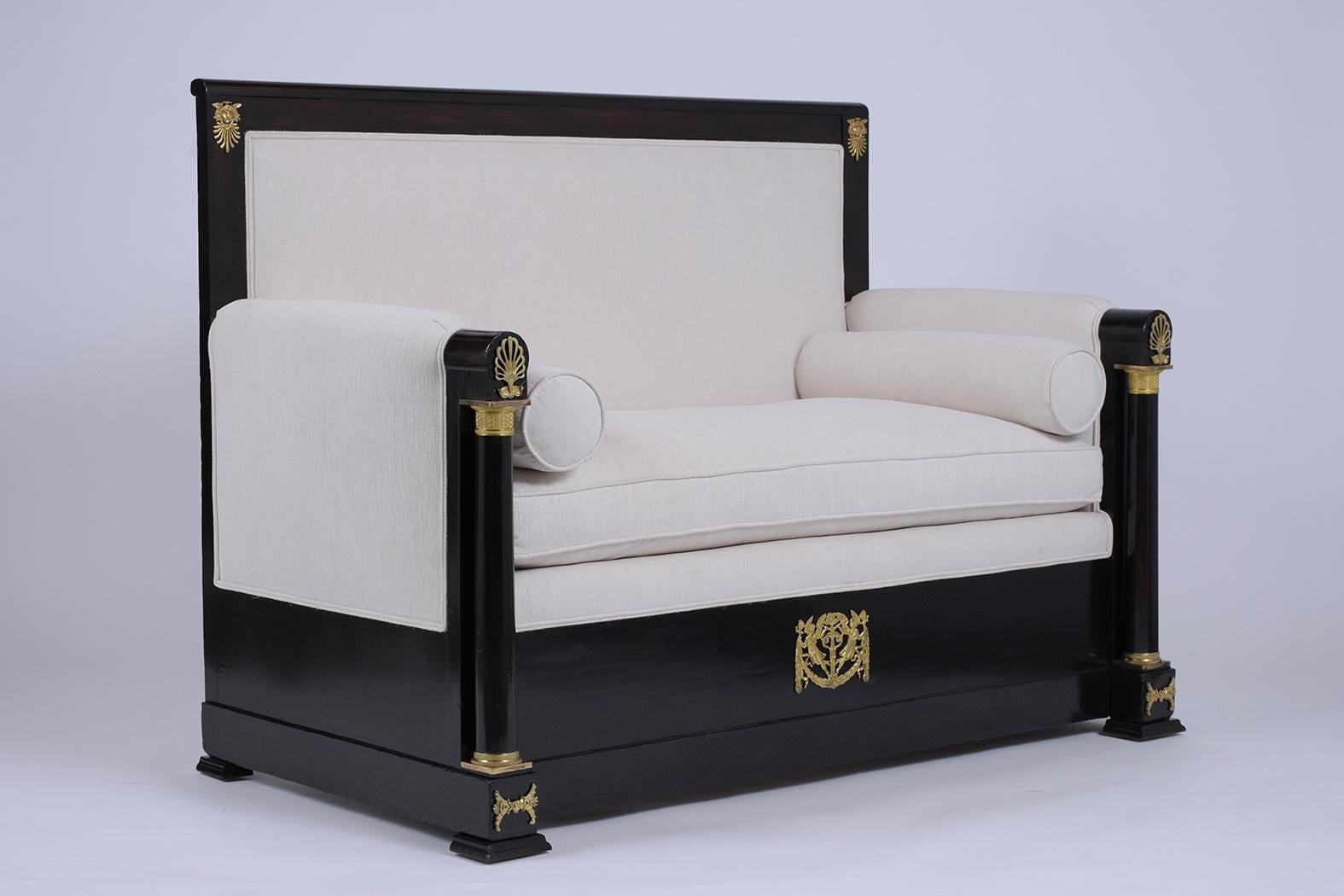 This Unique French Empire bench circa 1880s features a solid wood frame that has been stained in a rich ebonized color with lacquer finish. The frame features stunning ormolu accents on the top, sides, arm columns, and bottom. There are depictions