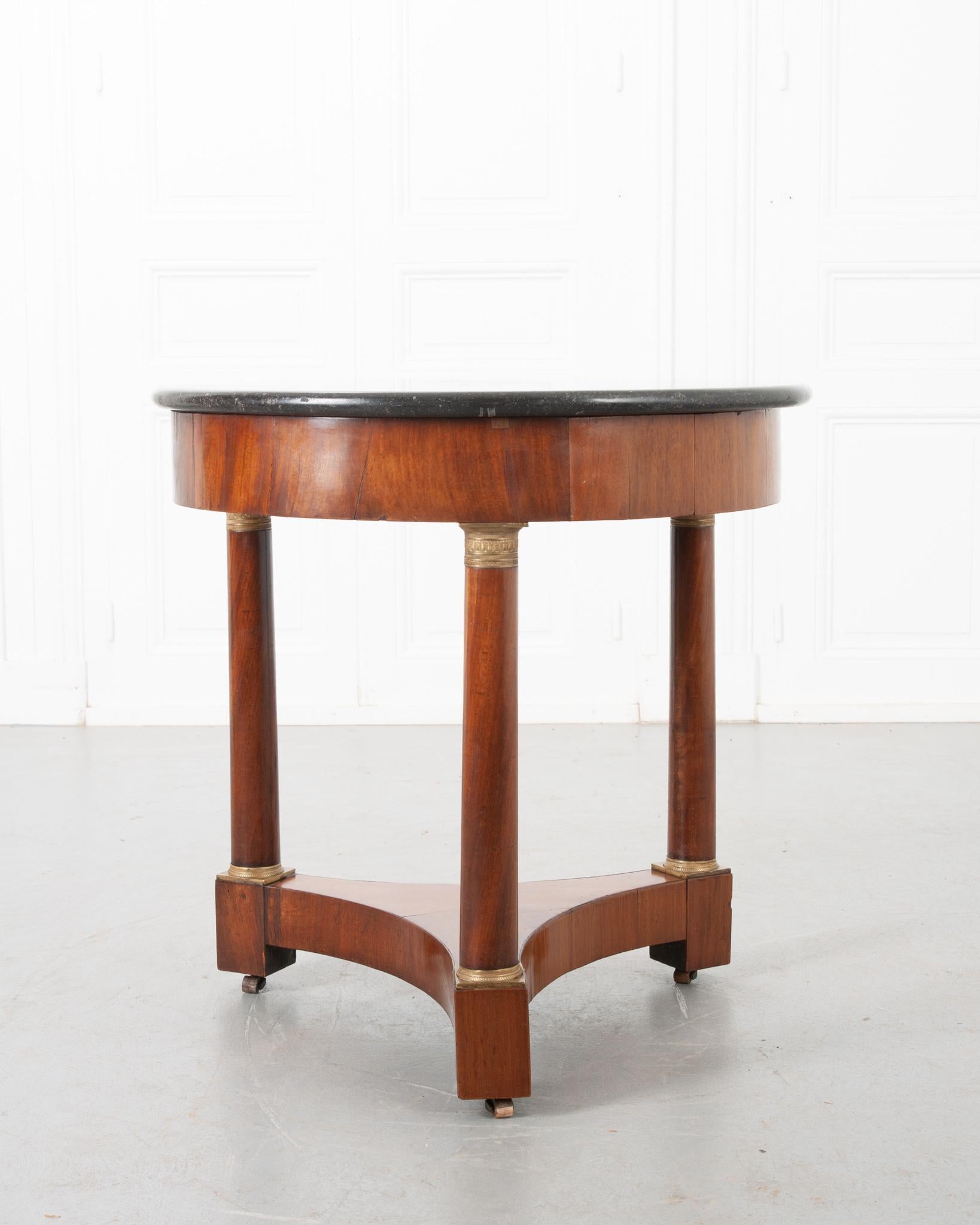 A darling Empire style gueridon table from 19th century France, with round black fossil marble over a simple mahogany apron. The three columnar legs have decorative brass ormolu capitals and bases, resting on a three-sided, concave shaped stretcher