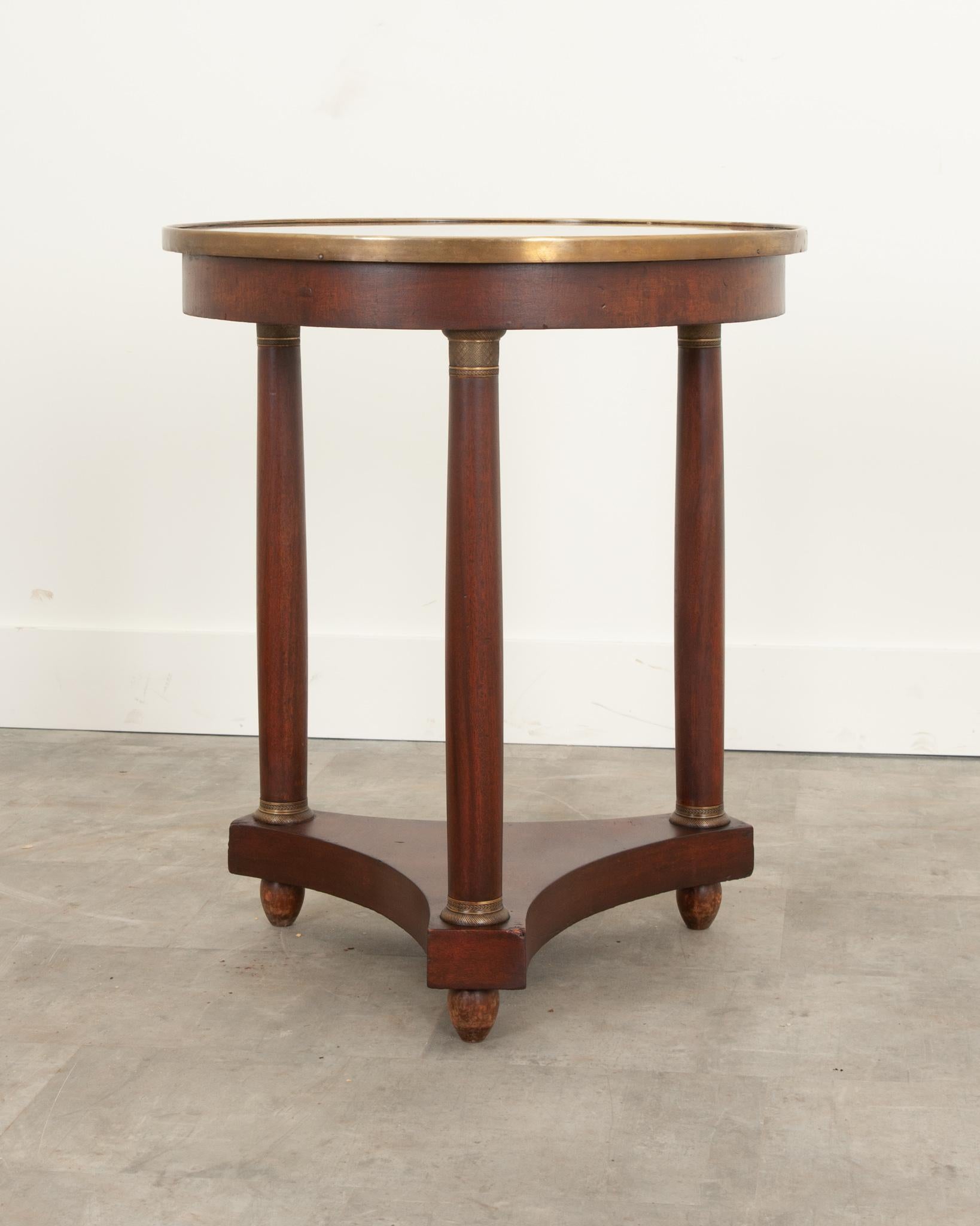 A handsome Empire gueridon crafted in France during the 19th century from beautifully patinated mahogany. Topped with black stone that’s in wonderful antique condition and encompassed with a thick, lustrous brass band. Three column-form legs feature