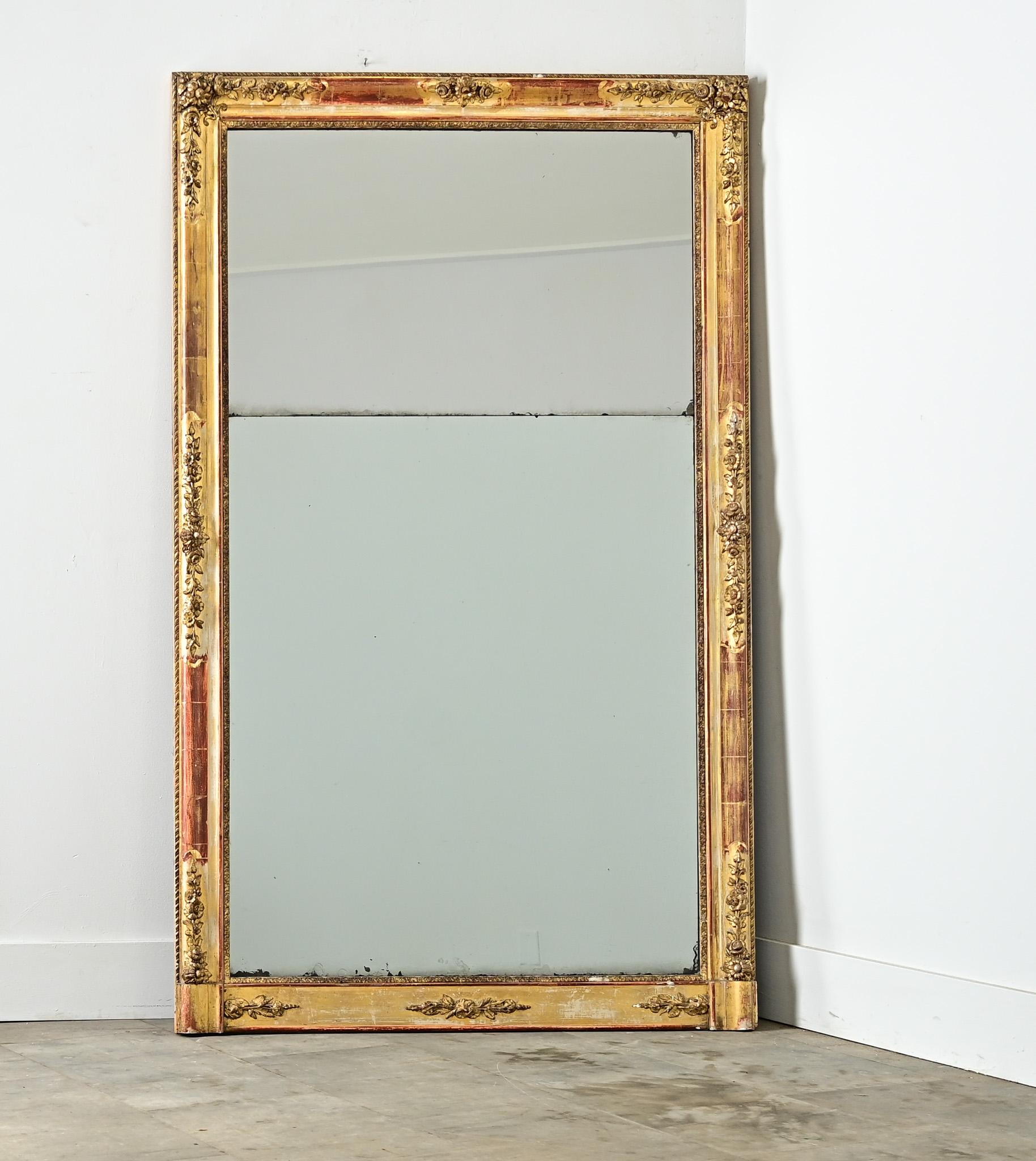 Questo specchio da camino dell'Impero francese (1800 circa) presenta una decorazione superficiale sobria, concentrata su motivi floreali intermittenti a basso rilievo che lasciano ampio spazio alla modanatura dorata per rivelare il suo bolo rosso