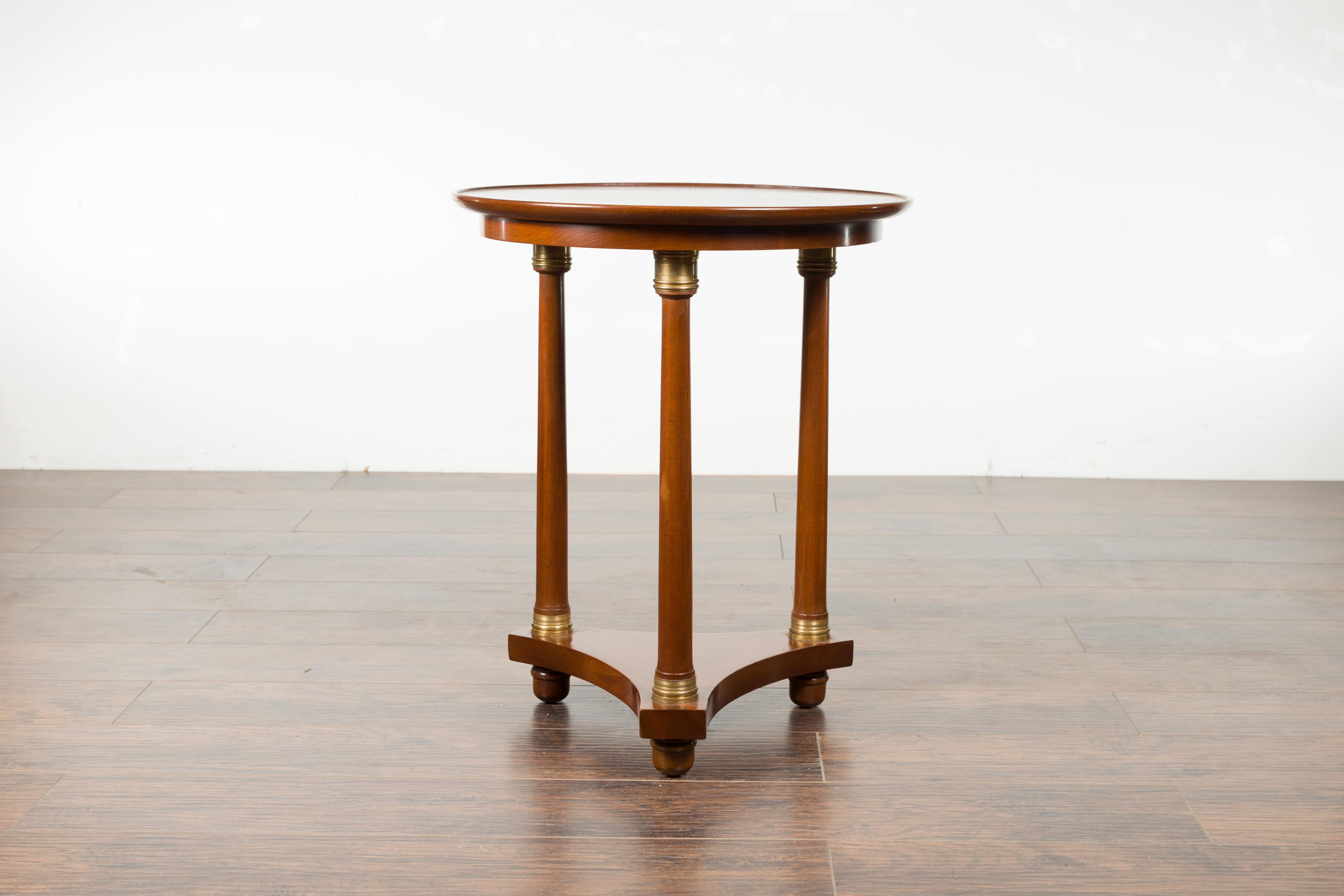 A French Empire side table from the 19th century, with bronze accents and lower shelf. Created in France during the politically dynamic 19th century, this Empire side table features a circular top sitting above three cylindrical legs adorned with