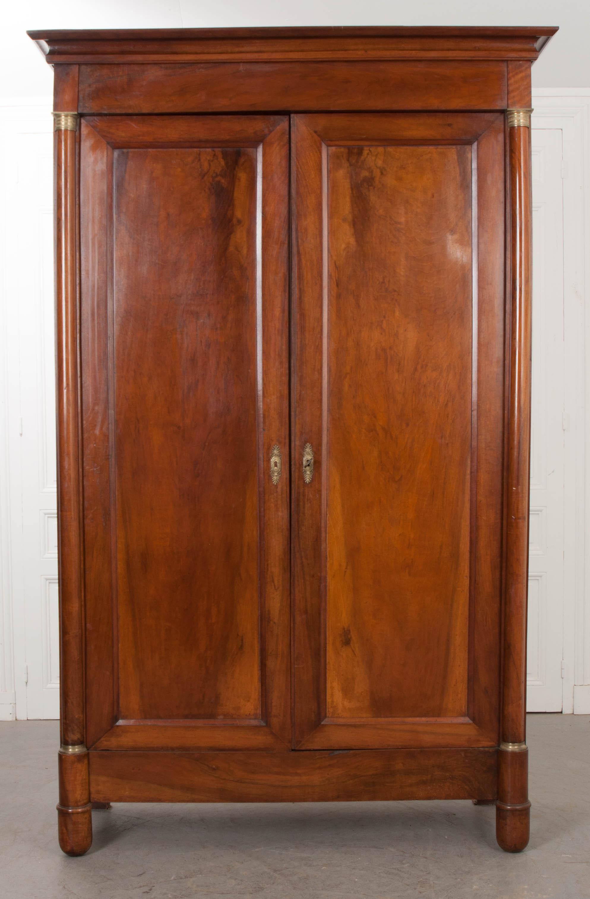 This elegant two-door walnut armoire is from France, circa 1850s, featuring a molded frieze over two doors with book-matched mahogany veneers and flanked by brass-collared pilasters. The doors, with brass palmette escutcheons, open to reveal two