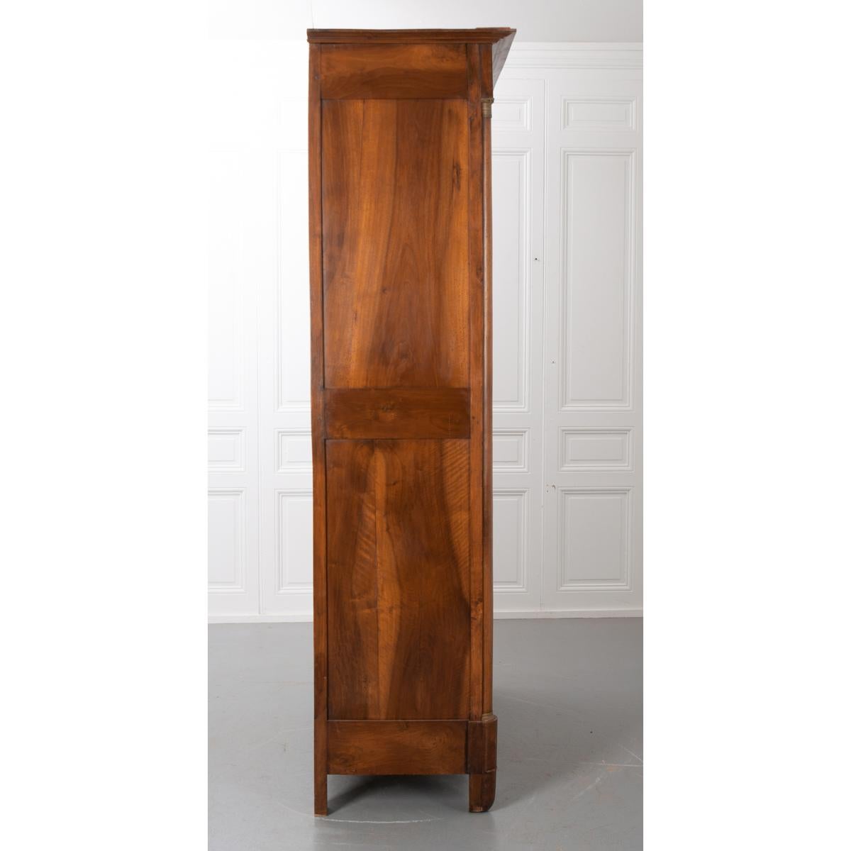 This elegant period Empire walnut armoire features a carved cornice over a book-matched walnut frieze, a pair of paneled doors mounted with stylized brass floral motif escutcheons. A single key works the right door and acts as a pull. The right door