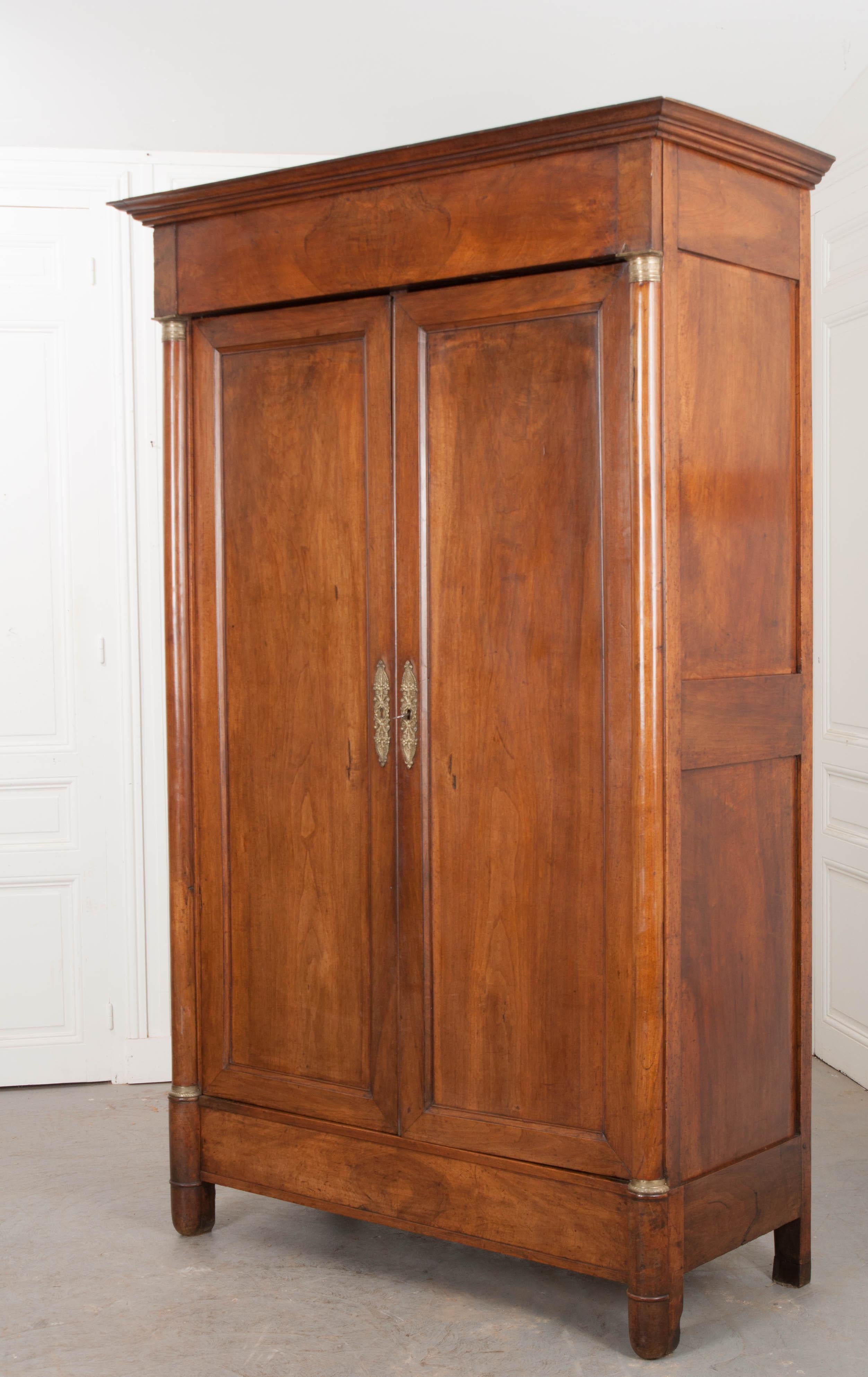 This elegant period Empire walnut armoire, circa 1830, is from France and features a carved cornice over a book-matched walnut frieze, a pair of paneled doors mounted with stylized brass escutcheons in “fruit-and-flower” decor and flanked by a pair