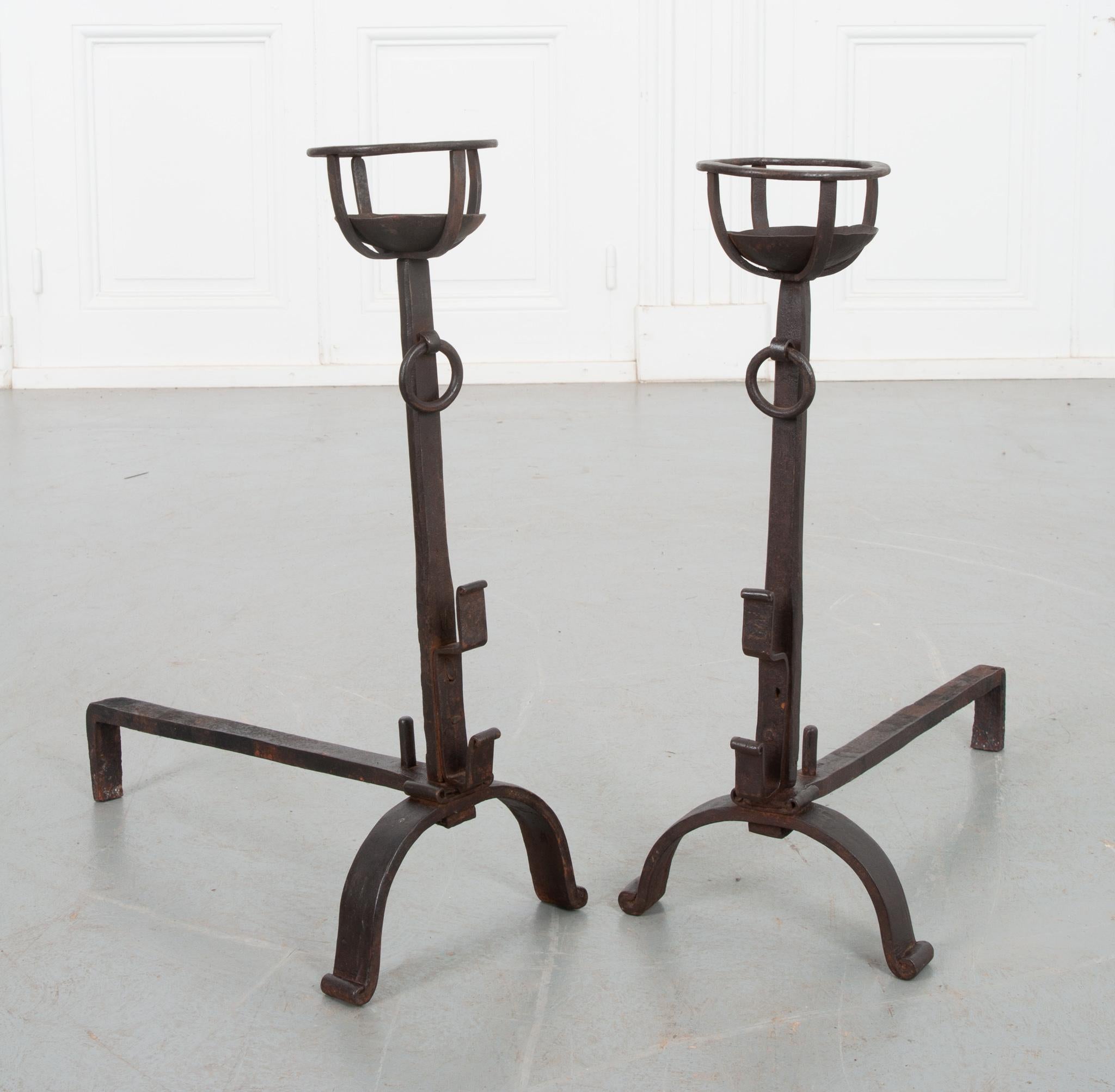 This pair of hand forged iron andirons comes from 19th century France. Used to support logs, these L shaped brackets are simply constructed. The tops are formed like open baskets above matching drop rings. A unique detail on the front of the