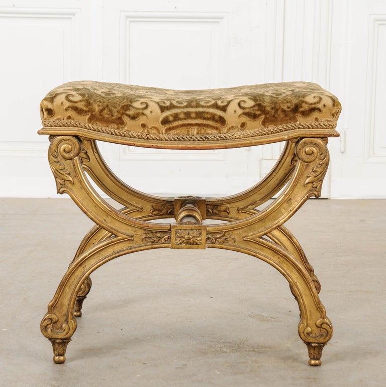A wonderful Empire style vanity bench with decorative X-form and turned wood stretcher. The upholstery may not be original, but is lovely and could be reused. The gilt frame is ornamented with foliage and rosettes and scrolled details, ending in