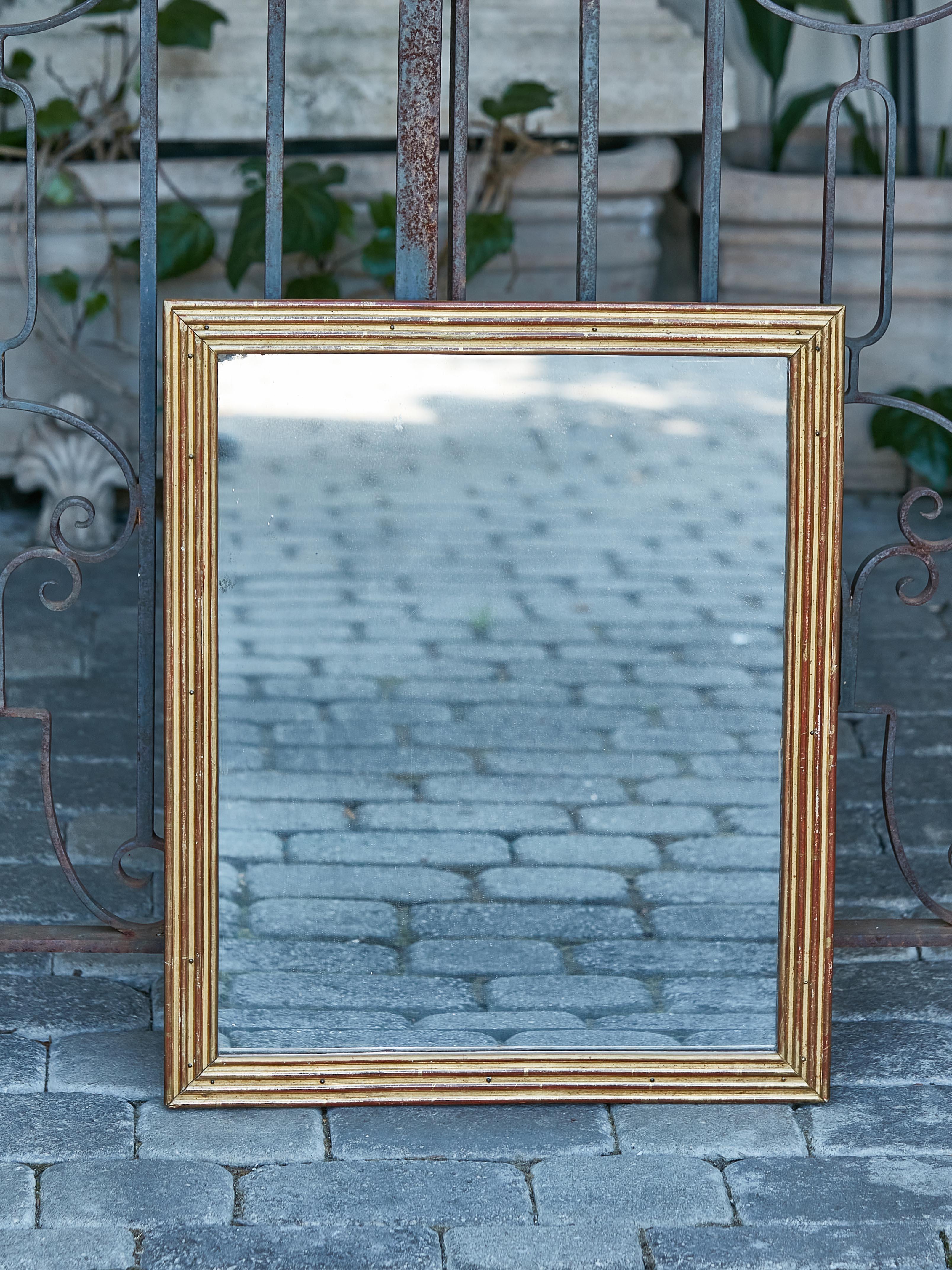 A French giltwood mirror from the 19th century with linear rectangular frame, reeded accents and red undertone. Behold the fusion of opulence and antiquity in this French giltwood mirror, a treasure dating back to the 19th century. Enveloped in a