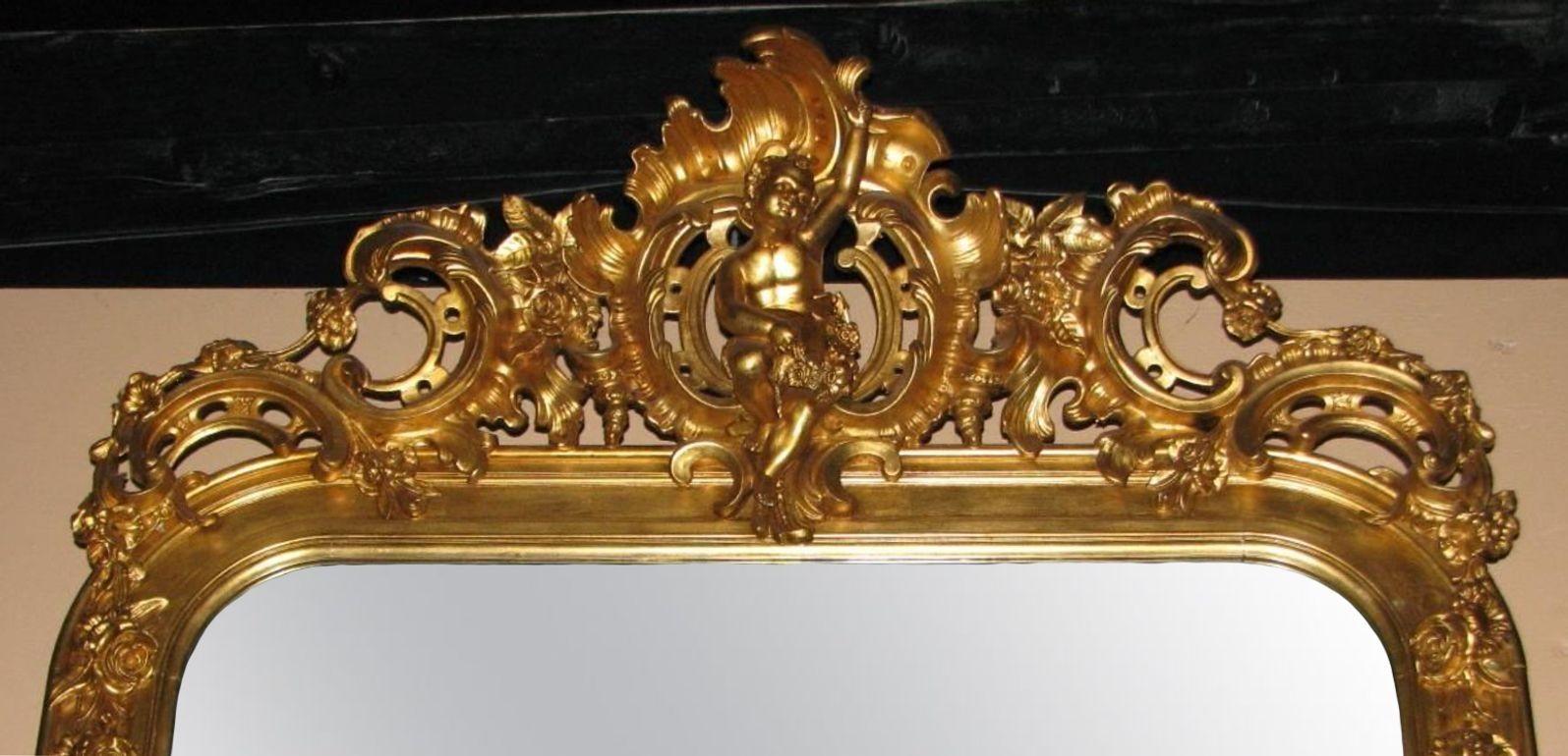 A French antique 19th Century hand-carved giltwood mirror having rocaille ornaments all around the frame, while also having a putti in the middle top holding a wreath. The piece includes two beautiful porcelain plaques, each depicting a romantic