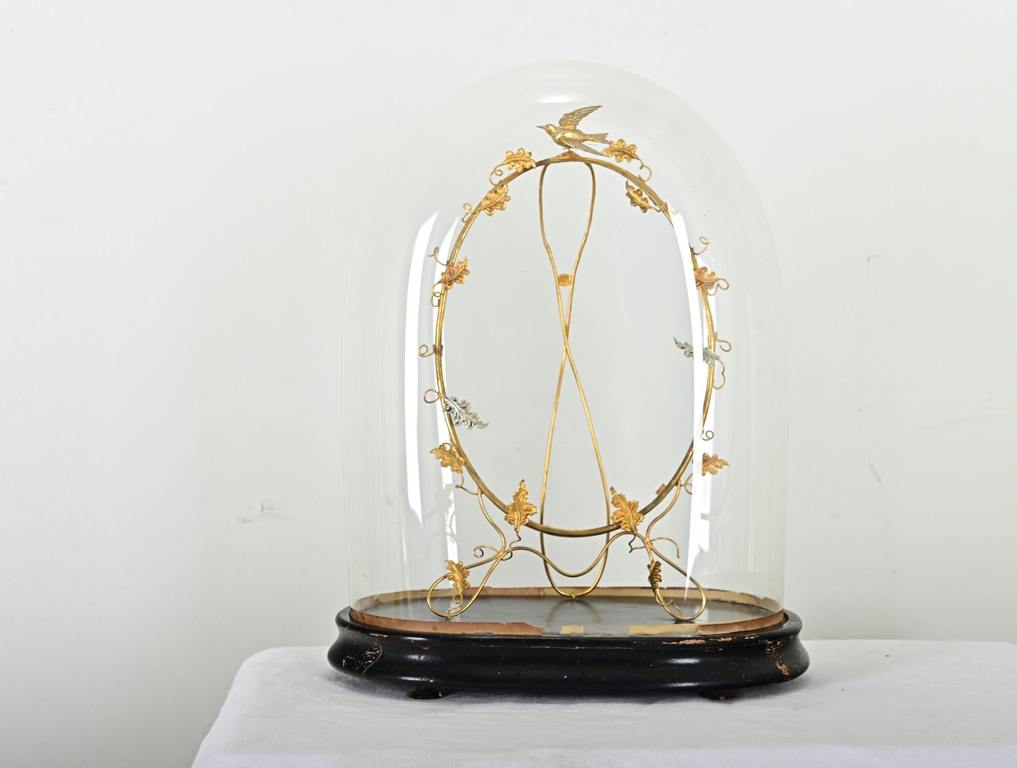 Popular in the 19th century, domed glass displays offered protection for portico clocks and flora & fauna specimens without compromising visibility. Inside this cloche is an oval easel frame, the wiry brass tendrils and charming bird figure