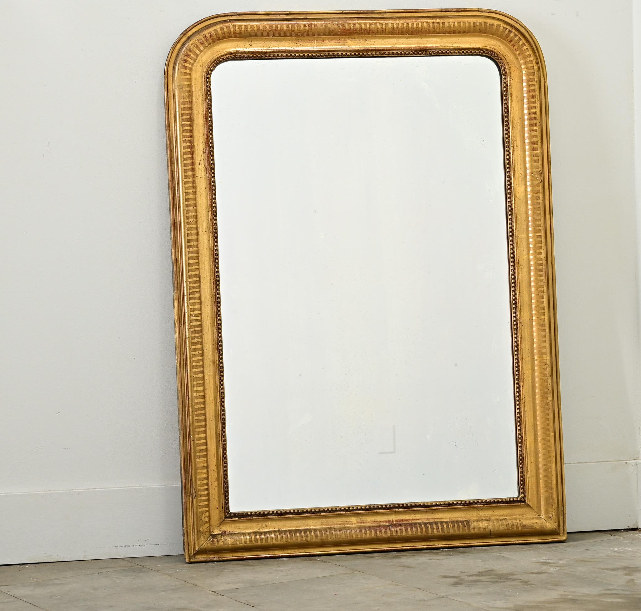 Miroir Louis Philippe du XIXe siècle, avec un cadre discret, doré, rehaussé d'une oxydation foncée et d'une texture craquelée. Le cadre est gravé d'un motif cannelé interrompu par quelques fissures et zones d'usure qui confèrent une patine