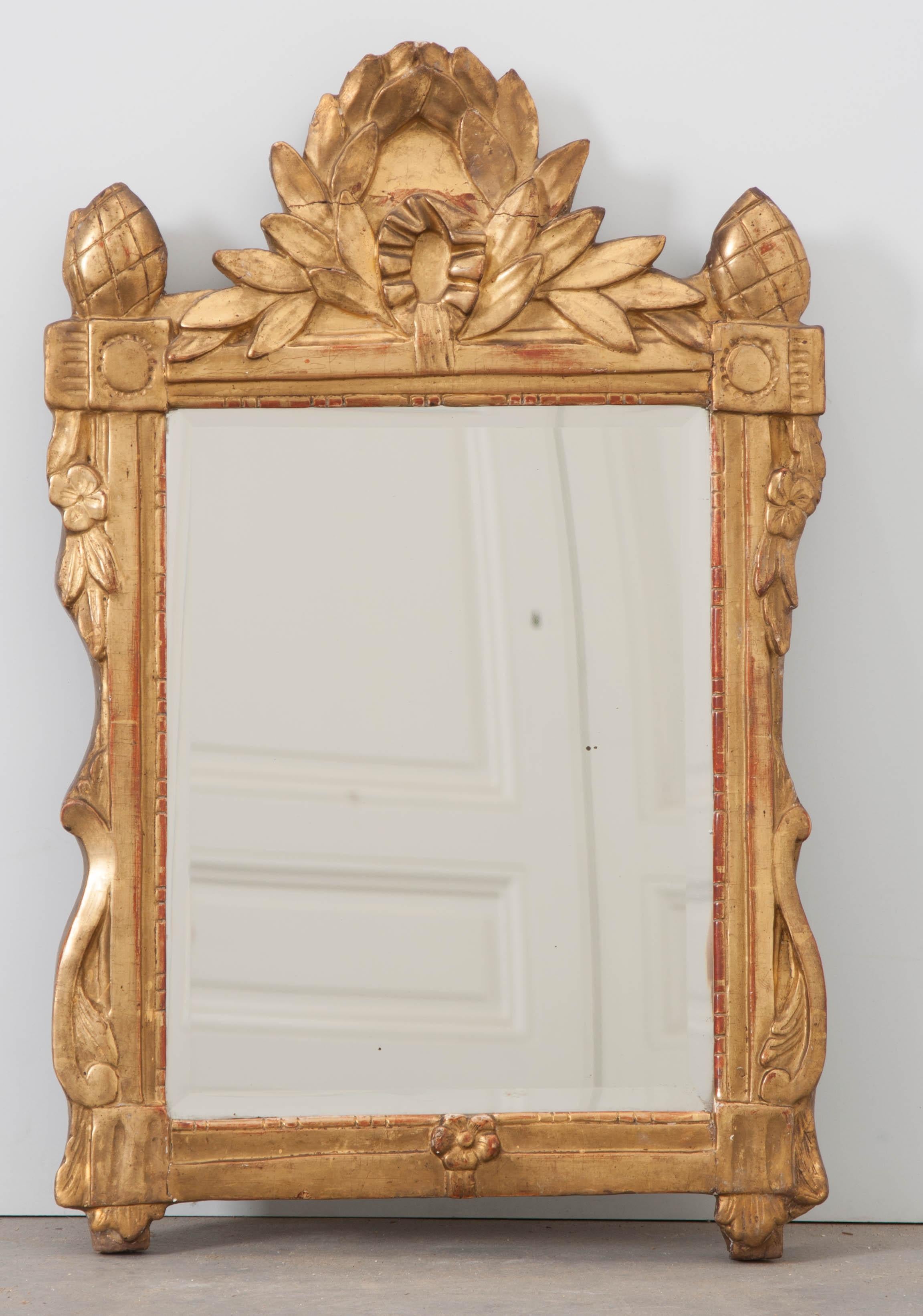 A brilliant Louis XVI style gold gilt mirror from 19th century France. Two gilt pineapples flank the central laurel wreath crest found atop the small mirror’s frame. Floral and scrolled details embellish the frame’s sides with a dashed beaded pearl