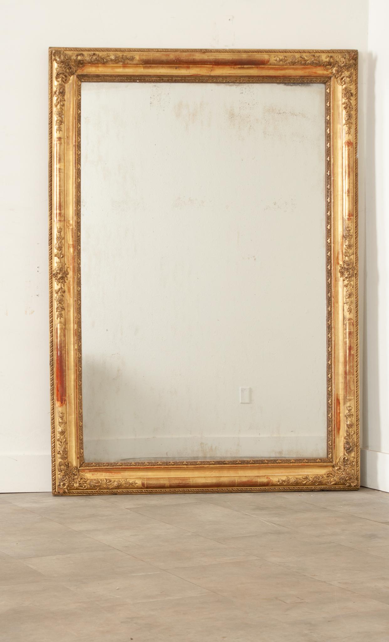 A French 19th century gold gilt framed mirror. The original mirror plate is secured in an intricately carved and molded frame on all four sides with repeating designs and floral accents. Some foxing is visible within the mercury mirror plate, adding