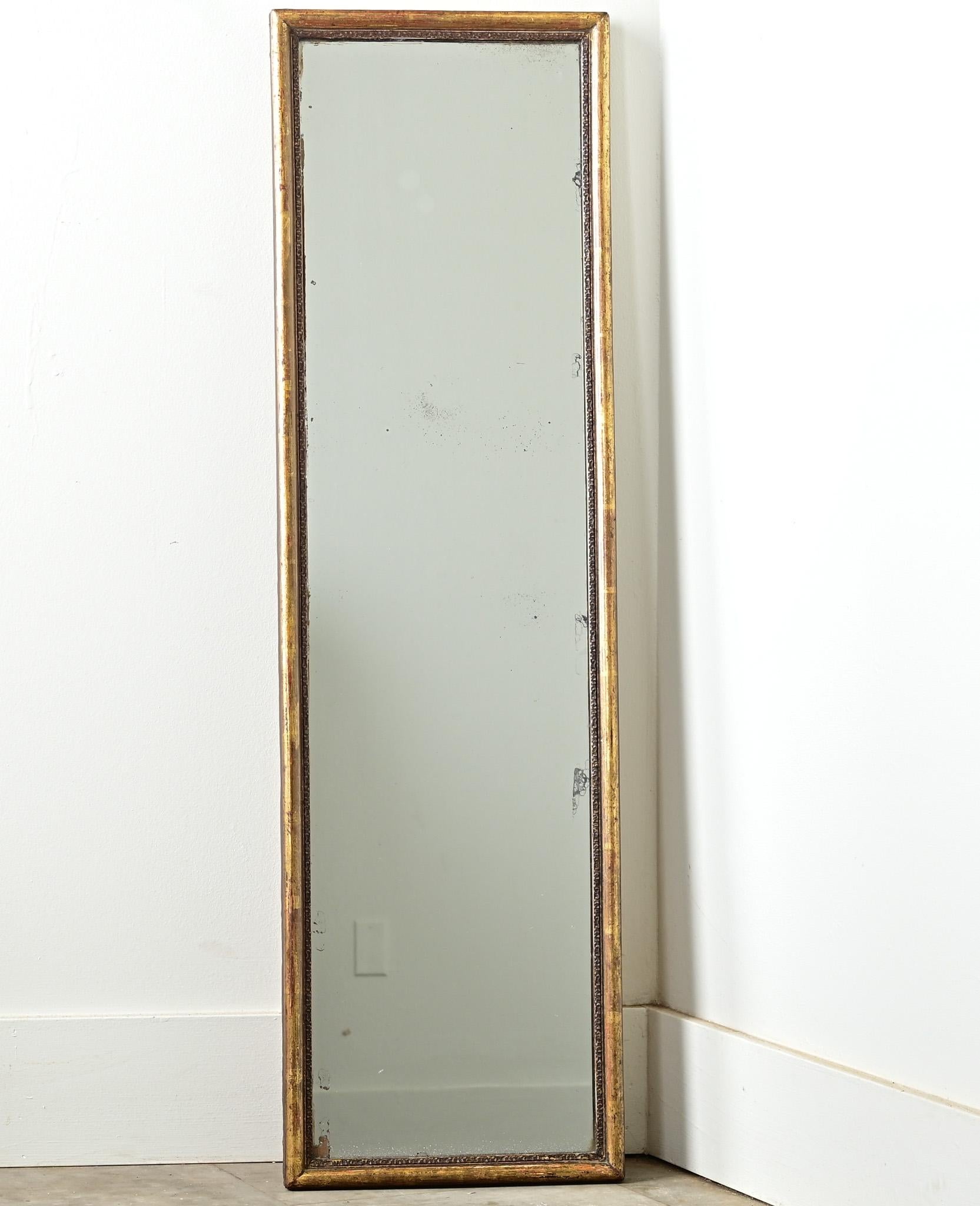 A narrow French gold gilt mirror from the 1800’s has its original gold gilt finish revealing the red bole underneath. The frame holds its original mirror plate with light foxing and crystallization settled at the bottom, and the original back