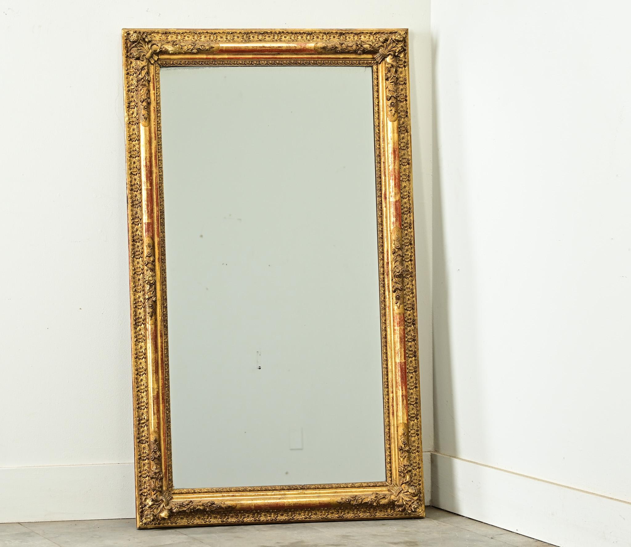A French gold gilt mirror from the 1800s in narrow proportions, though dense with luxurious floral carvings on each corner and in parallel rows of molding around the rectangular frame. This nearly symmetrical frame has a lustrous gilded finish