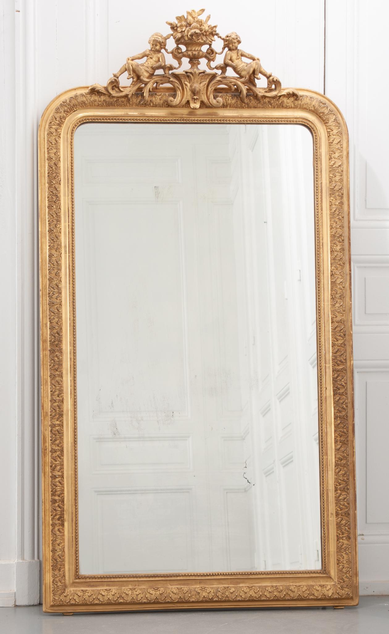 This is a French 19th century Louis Philippe style gold gilt mirror with a crest. It has its original mirror plate with some aging and foxing. There is a leaf motif all around the border. The crest has two cherubs, one on either side of a basket of