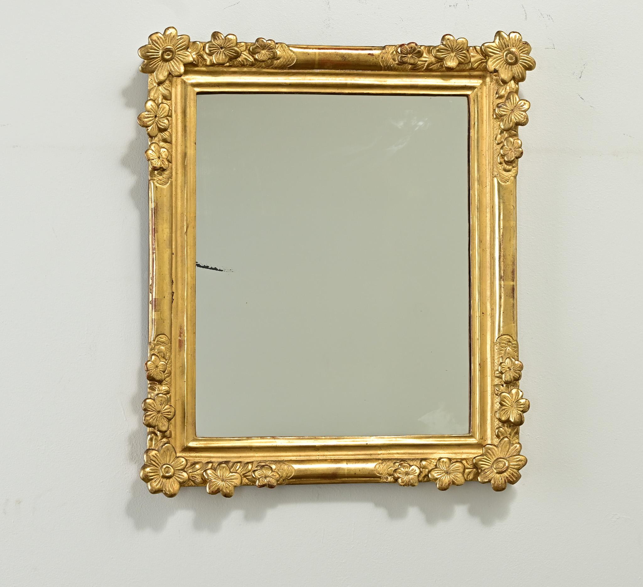 A petite French gold gilt mirror from the 1800’s is hand carved with impressive floral carvings in all four corners. This symmetrical frame has a lustrous gilded finish, and holds its original mirror with light foxing and sparkles that have settled