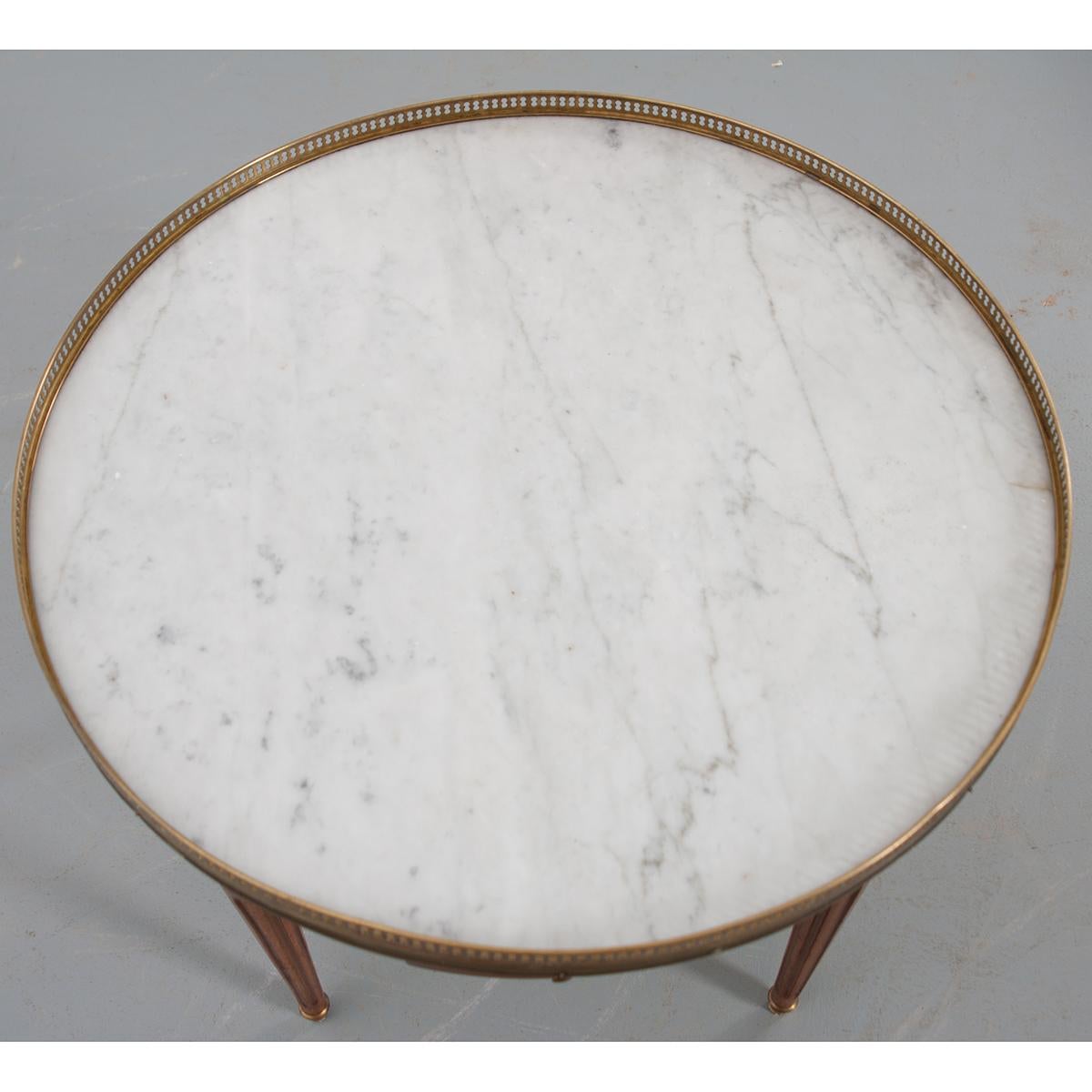 A classic example of a Louis XVI style marble-top guéridon coffee table, with brass gallery. The white marble top with reticulated brass gallery rests over an apron having two drawers and two green leather-topped slides, all with brass pulls. Fluted