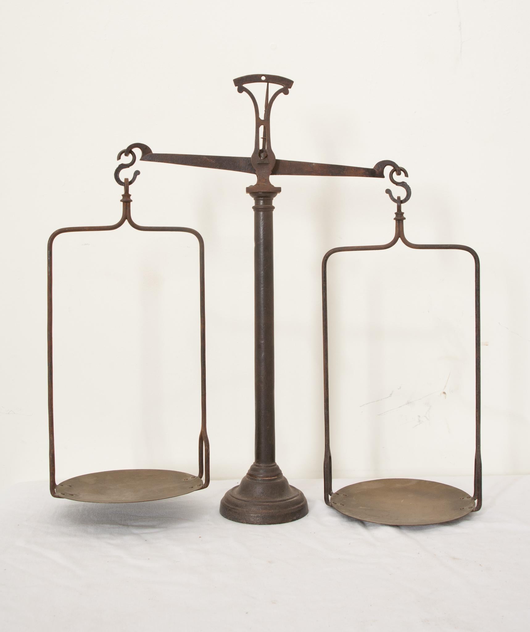 A smart French metal hanging balance scale from the 1900’s. Two straight stirrups hang from the arms with round brass dishes that hold the weighted item in one, and counterweights in the other. The scale has a column shaft, with a circular plinth