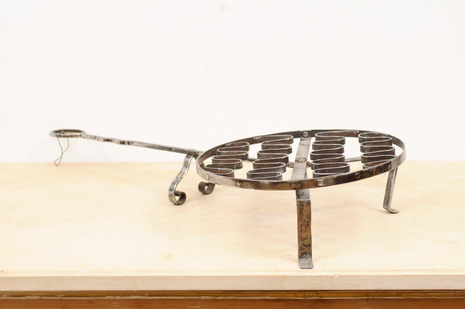 A French iron grill from the 19th century, with petite feet and long handle. Created in France during the 19th century, this iron grill was used as a tool in a fireplace. Featuring a circular body resting on petite feet, the grill showcases a long