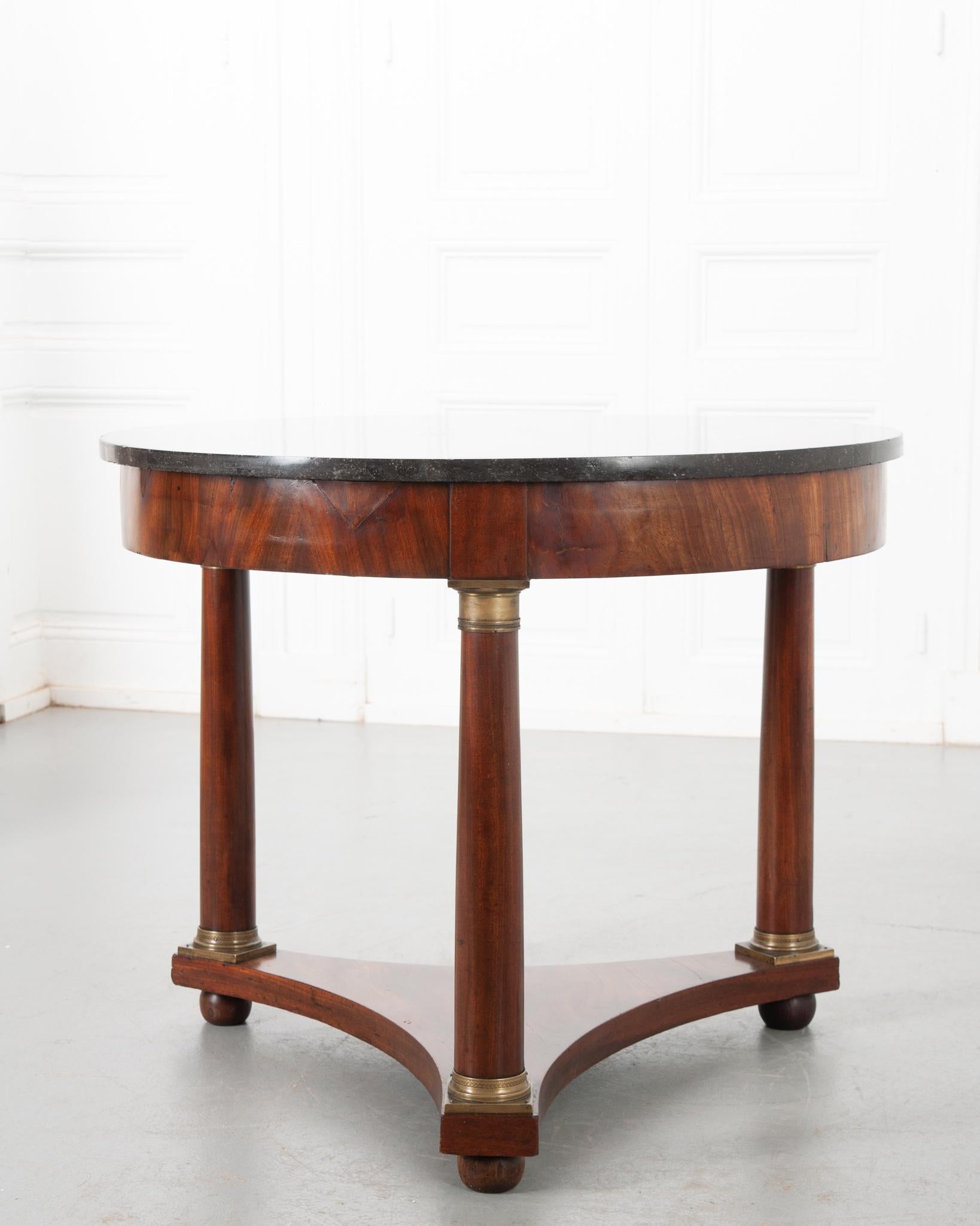 A sizable Empire style gueridon table from 19th century France. Beautiful black fossil marble over a mahogany body. A simple apron showcases the deep toned wood. Three columnar legs with decorative brass ormolu capitals and bases rest on a