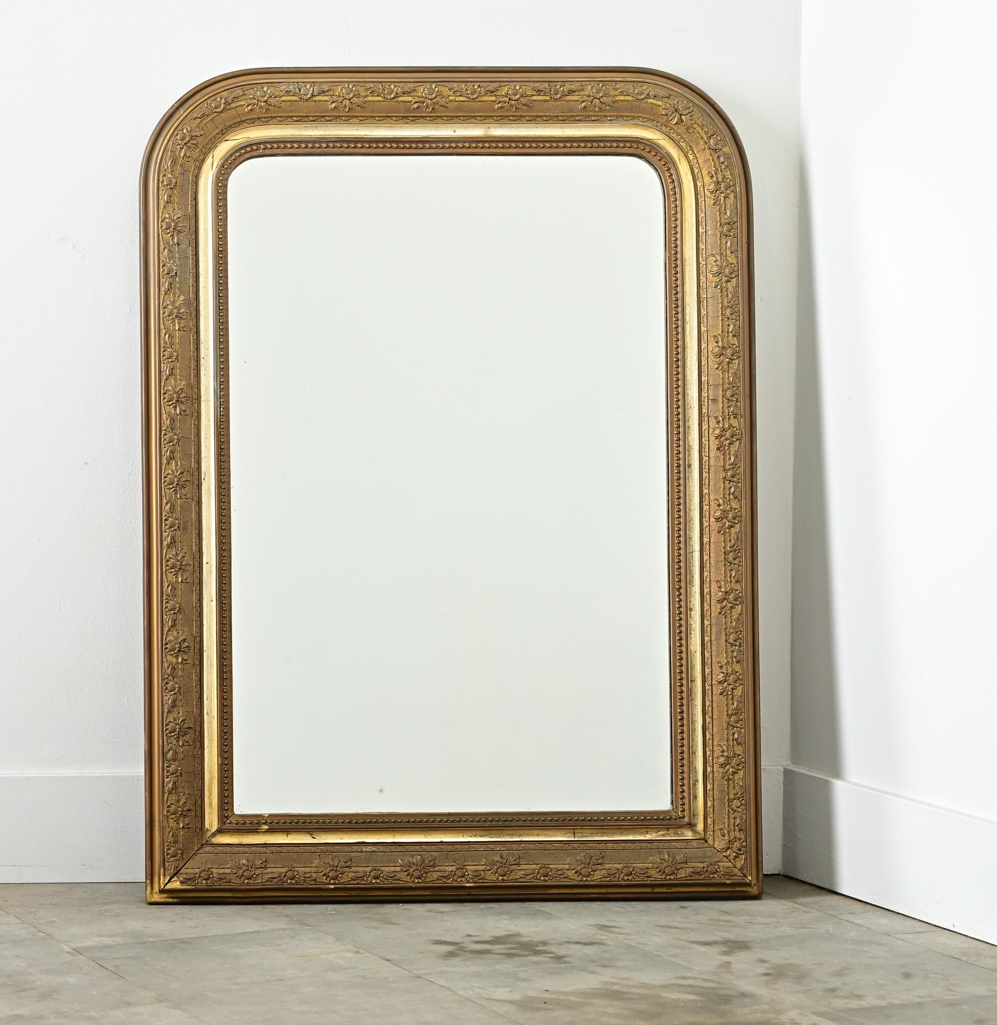 Intricate patterns of floral vines trace the wide molding on this 19th century Louis Philippe gilt mirror in classic, arched silhouette. The frame exhibits fine craquelure texture and antique-gold patina with a low coin-edge pattern toward the