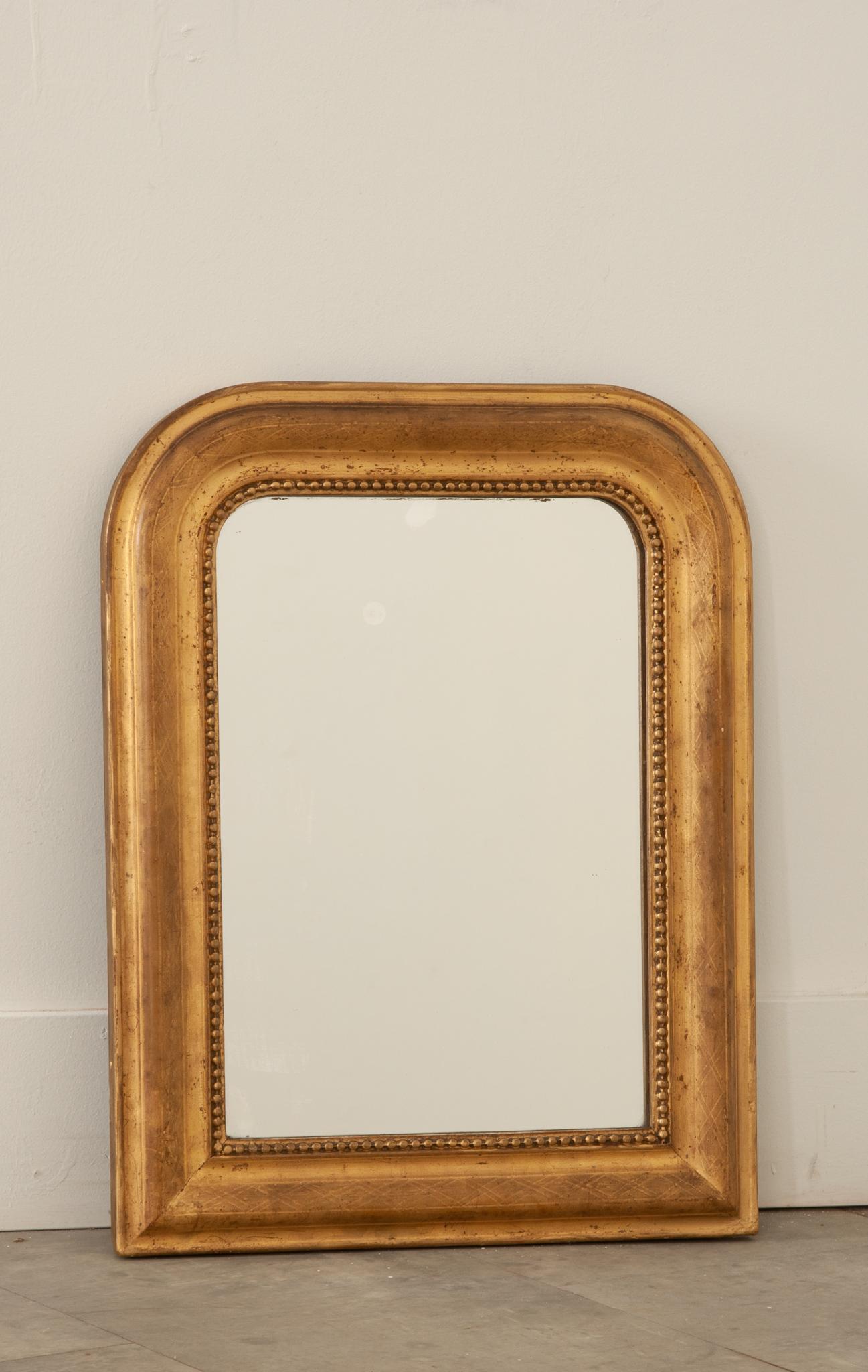 A darling petite mirror crafted in 19th century France. The original mercury mirror is in great condition and is surrounded by a carved bead border. A faint crosshatch pattern decorates the frame. The quality gilt has gained a beautiful patina over