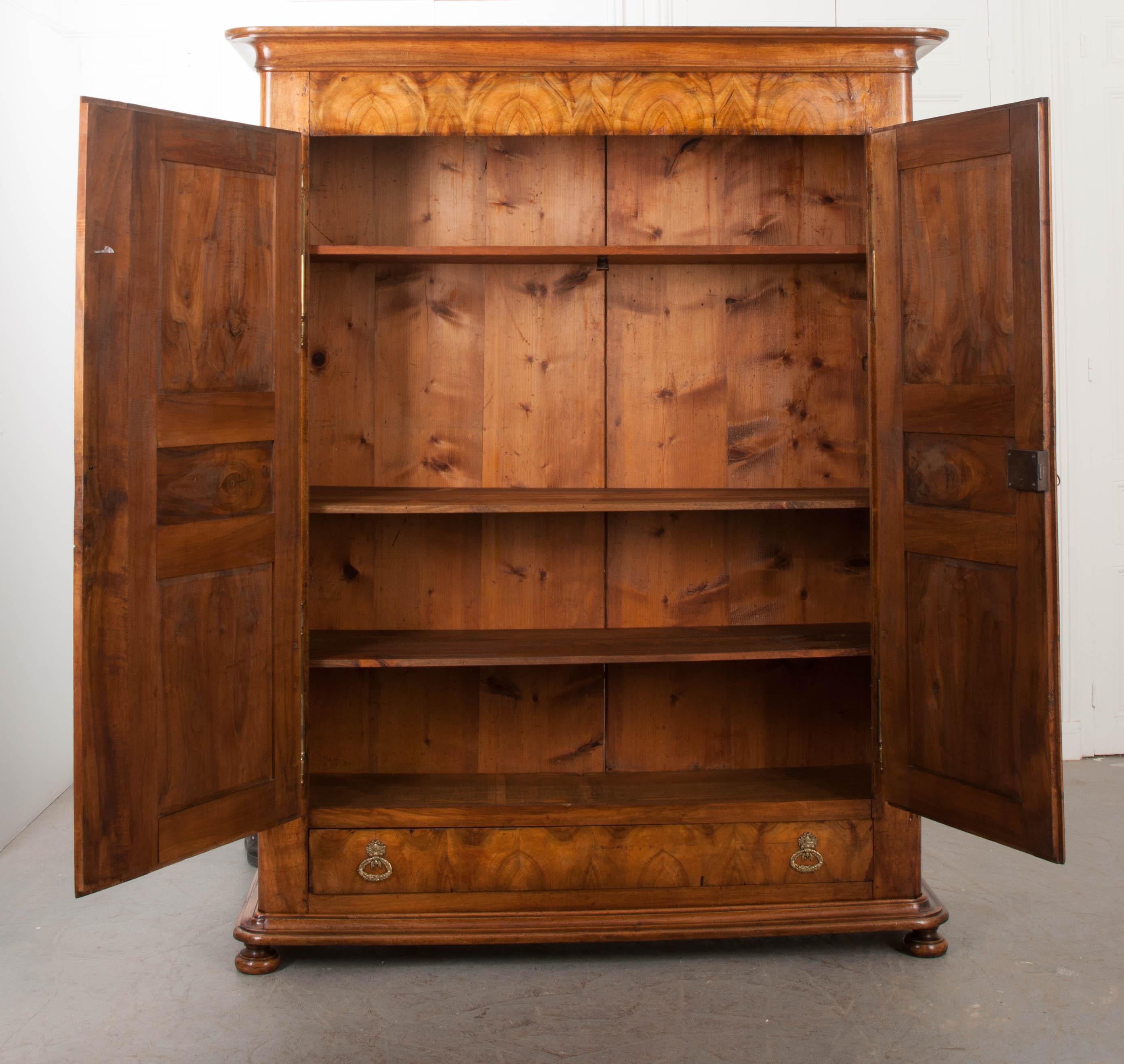 This stunning 19th century walnut armoire, circa 1850, is from France and features a carved crown with rounded corners, over a frieze with gorgeous bookmatched walnut veneers. The two tri-paneled doors open to reveal three removable shelves over a