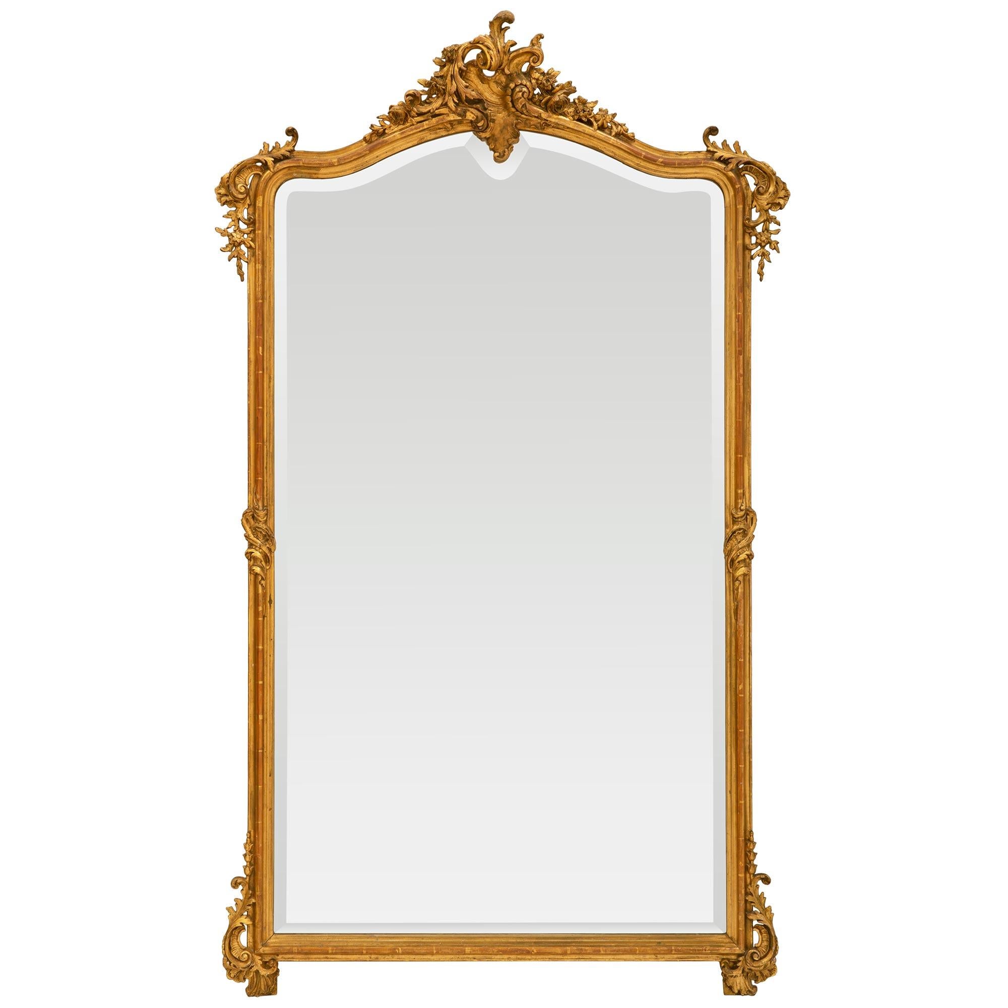 A large and finely carved French 19th Century Louis XV st. giltwood mirror. The mirror with all of its original gilt and beveled mirror plate has carvings of a central top reserve with scrolls and each side floral garlands. On all four corners of