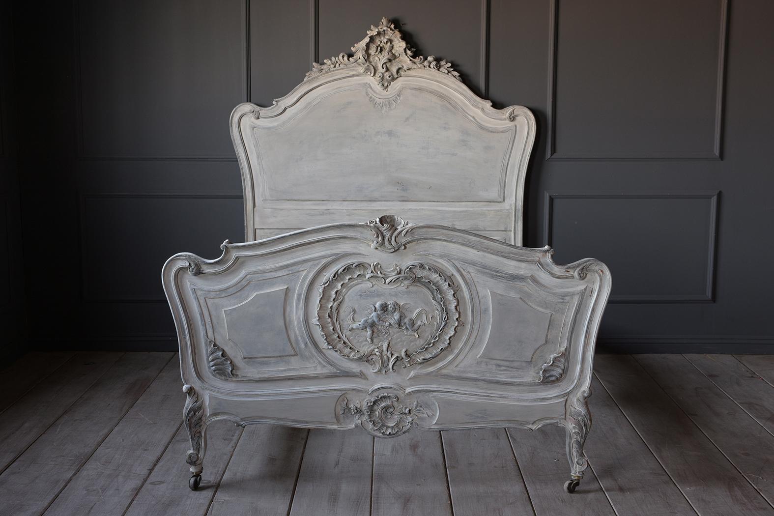 This French 19th century Louis XV style bed frame fits an XL twin mattress. It has a solid wood frame that is painted a pale light blue, and grey with a distressed finish. It has carved details on the headboard, footboard, and wooden rails. The legs