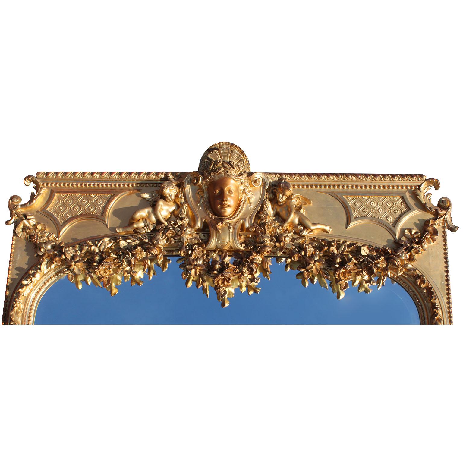 A very fine and palatial French 19th century Louis XV style carved giltwood and gesso figural beveled mirror frame trumeau. The arched plate within a main foliate and acorn trailing border, surmounted by ribbon tied floral swags centered by a mask
