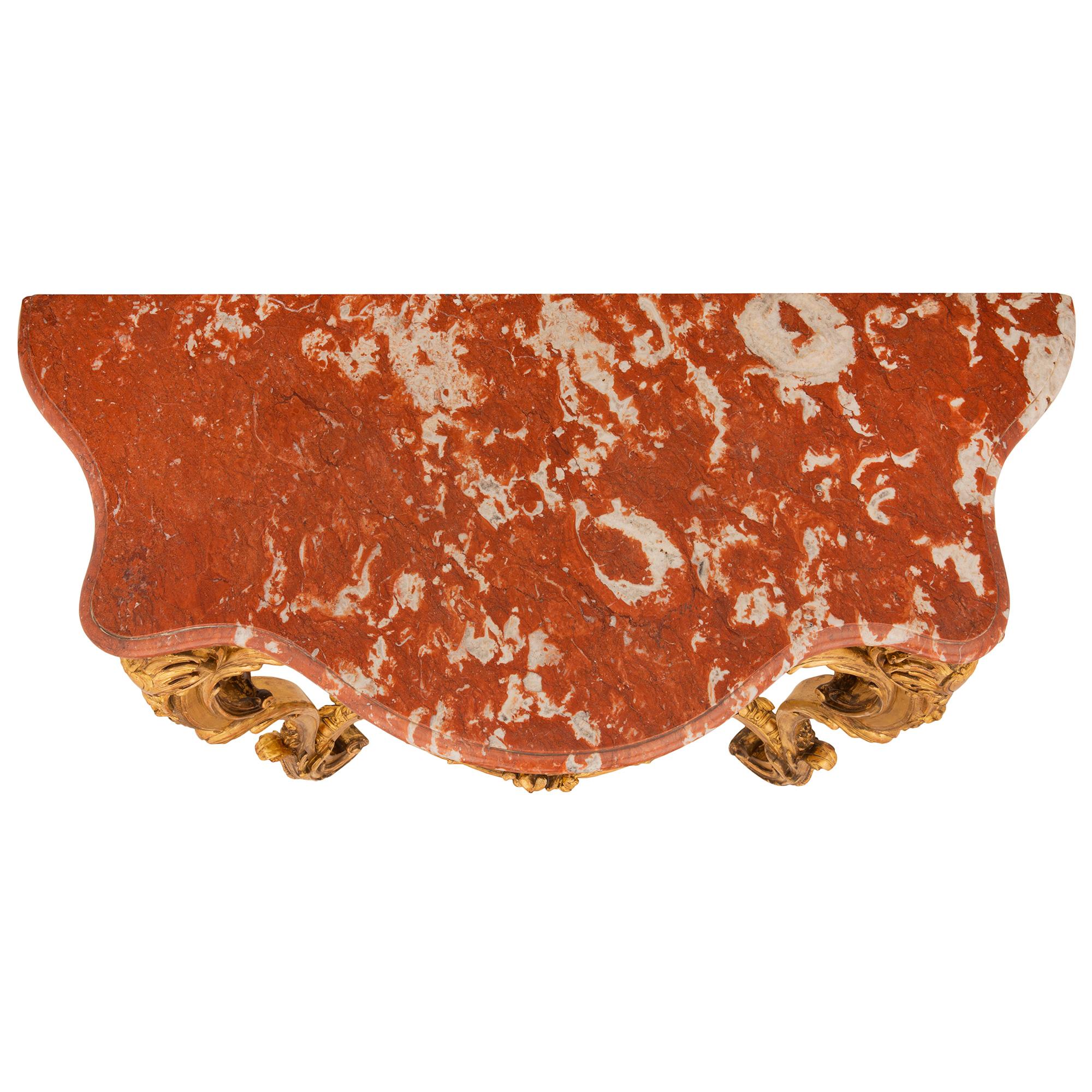Elegantísima consola francesa del siglo XIX de madera dorada estilo Luis XV y mármol Rouge Royale. La consola se eleva sobre hermosas patas con roleos y movimientos foliados ricamente tallados, hojas de acanto y un fino entramado. Las patas están
