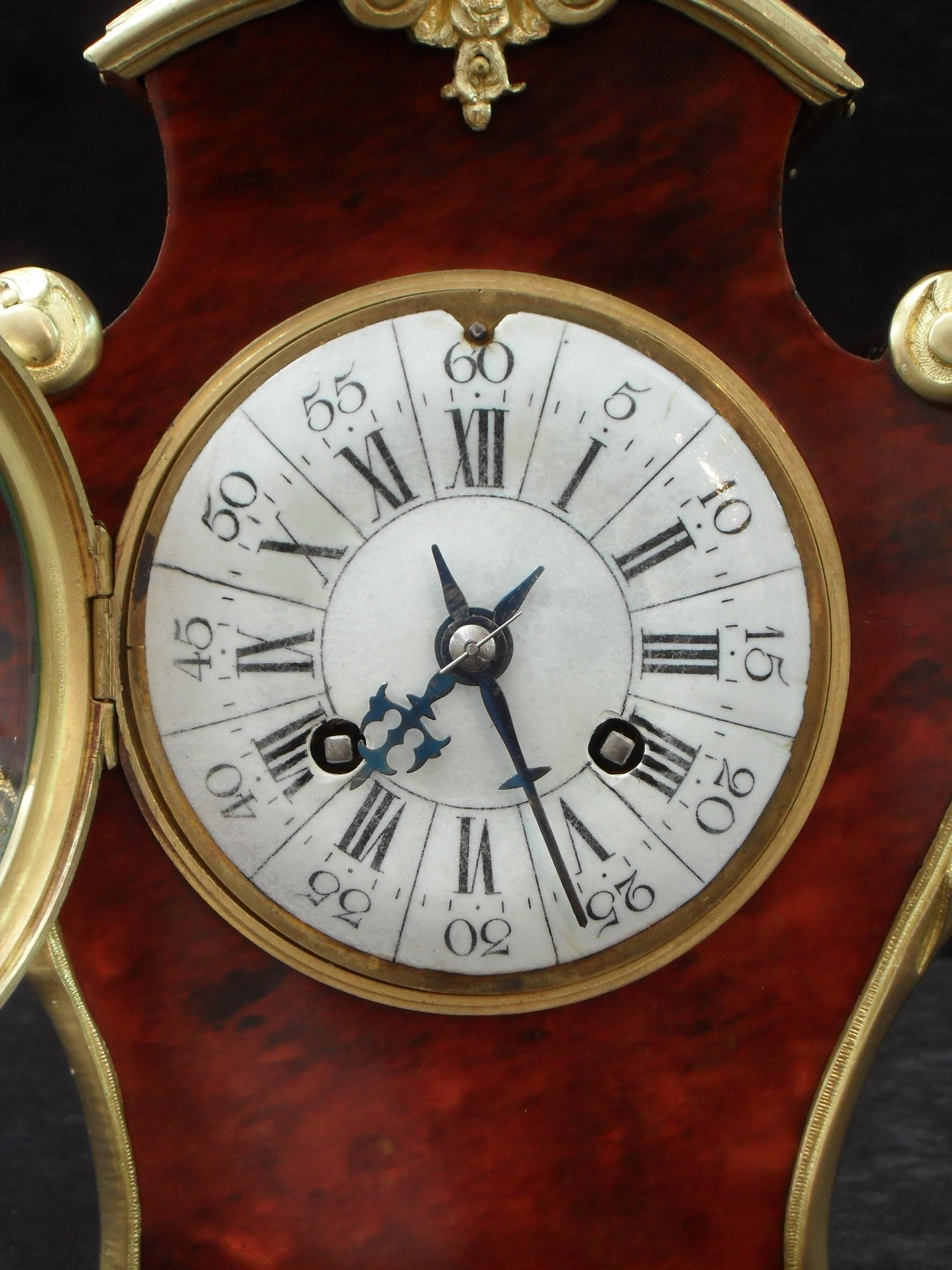 A very good quality French Louis XV style red tortoise shell serpentine shaped mantel clock with scroll leaf bronze gilt ormolu mounts and carrying handle. The clock has a white enamel dial with a French eight day movement which strikes the hours