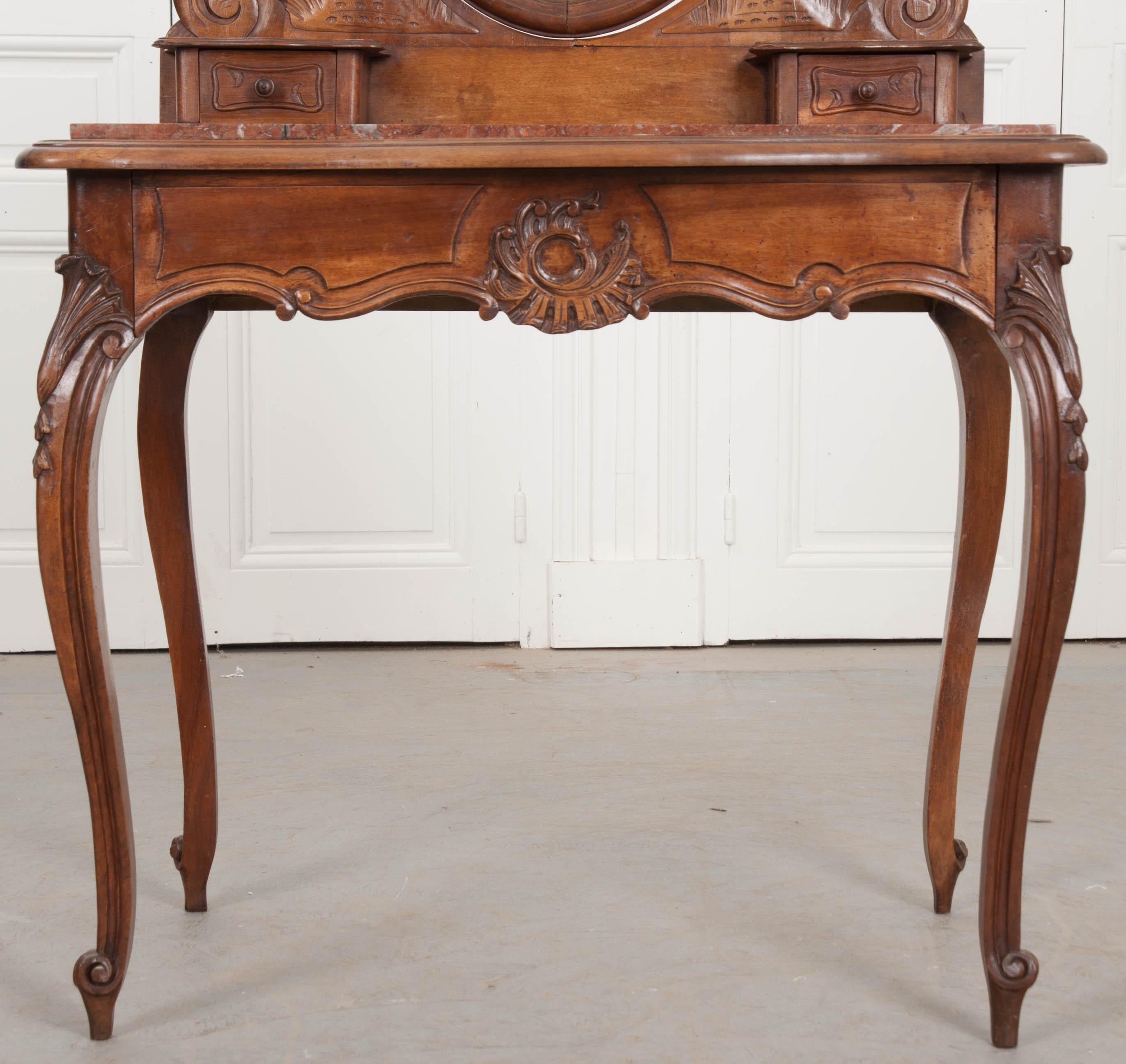 An expertly carved walnut vanity, done in the Louis XV style, circa 1890. The dressing table features a tilting mirror, three-drawers, and an inset marble top. The marble top is red in color, with black, gray and white veins running throughout. The