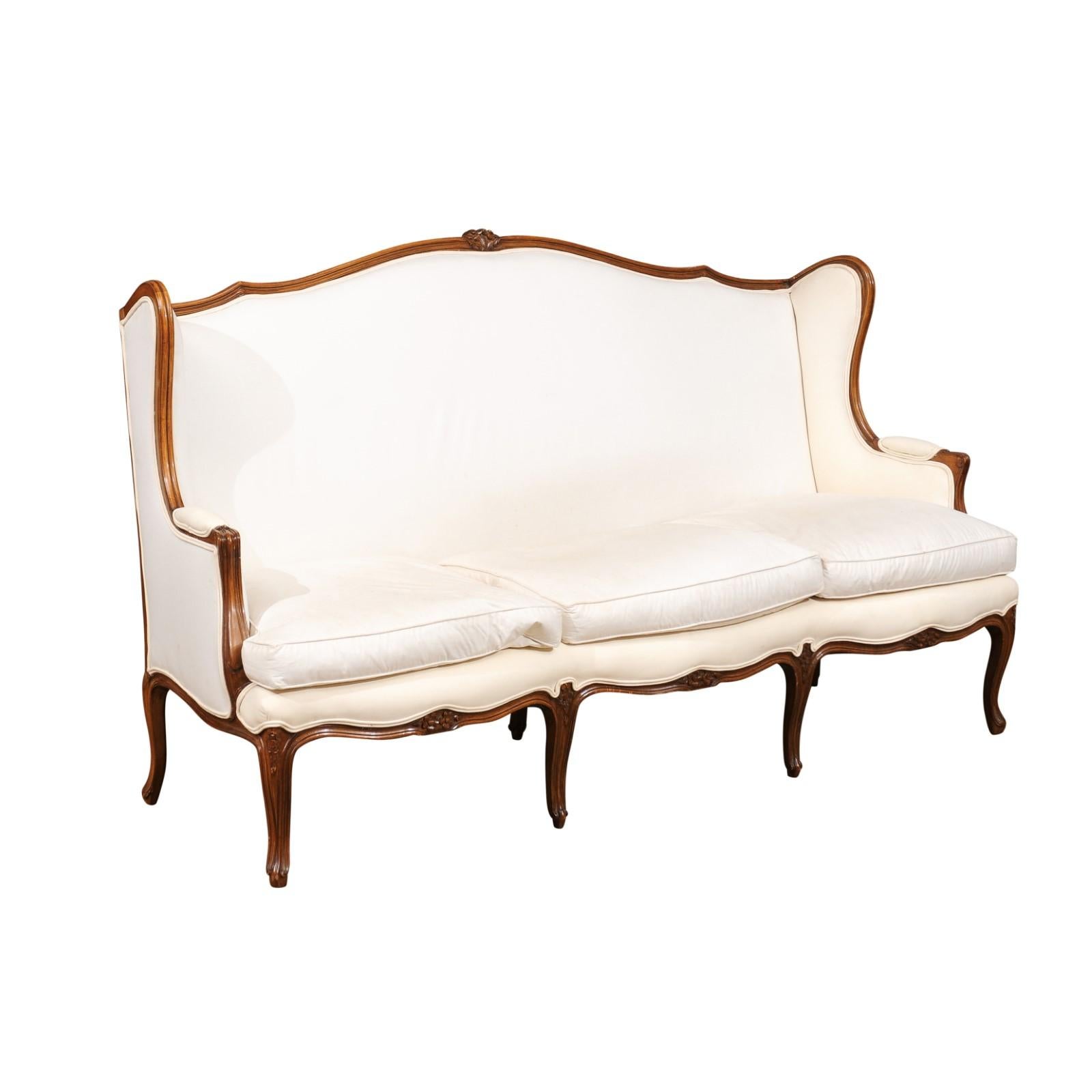 A French Louis XV style three-seat wooden winged sofa from the 19th century with carved crest, scrolled arms, cabriole legs and new upholstery. This French wooden canapé à oreilles features a delicately curved upper rail accented in its center with