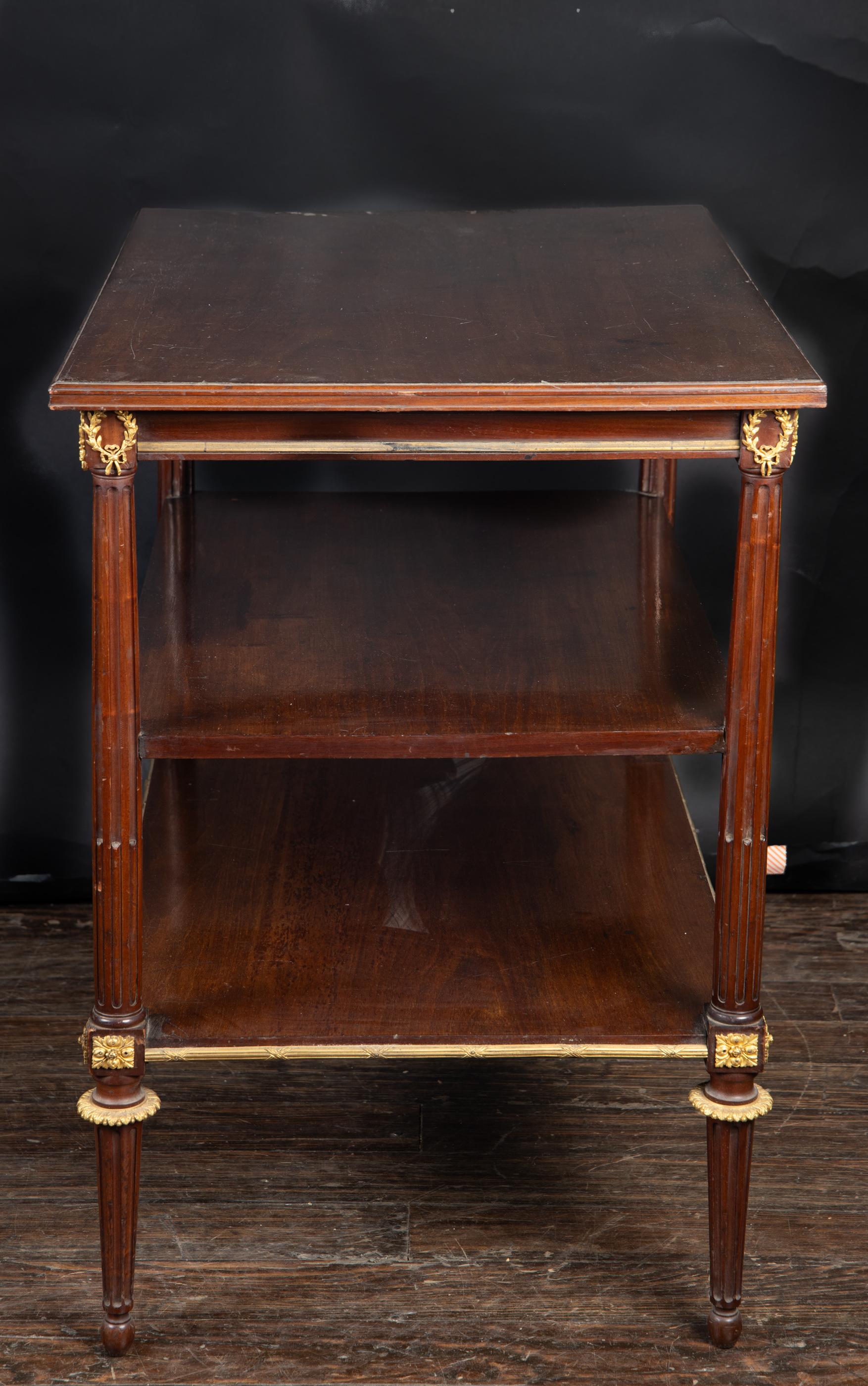Esta hermosa mesa de caoba de tres niveles presenta monturas de bronce diseñadas a la manera Luis XVI, y se apoya en cuatro patas de madera estriadas. Esta pieza antigua francesa, que data del siglo XIX, tiene una parte superior rectangular envuelta