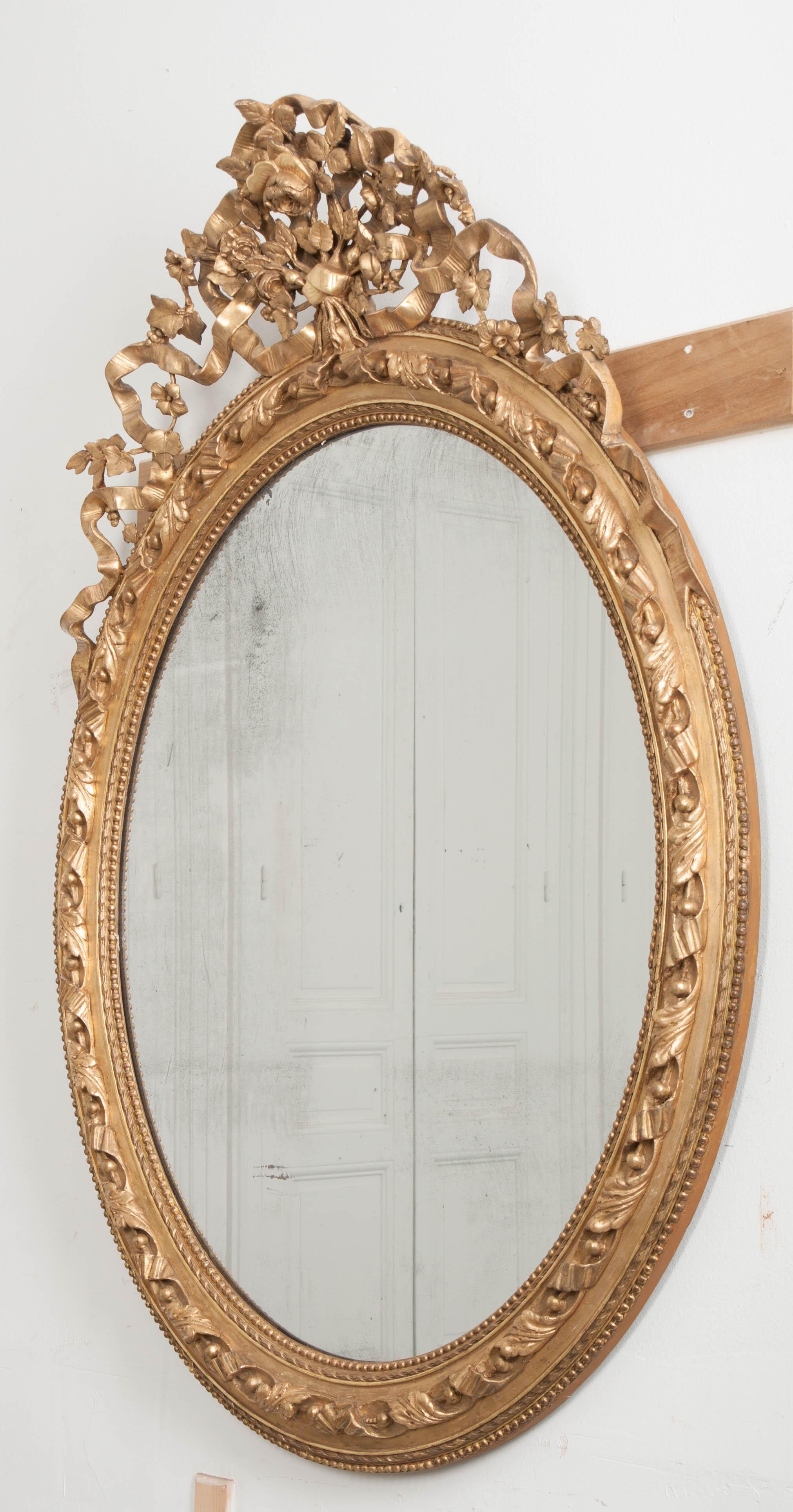 Fanciful and fun, this 19th century French Louis XVI- style oval gold gilt mirror boasts intricate, carved details from top to bottom. A carved bouquet of flowers and vines crowns the brilliant mirror, with exceptional ribbon details. The original