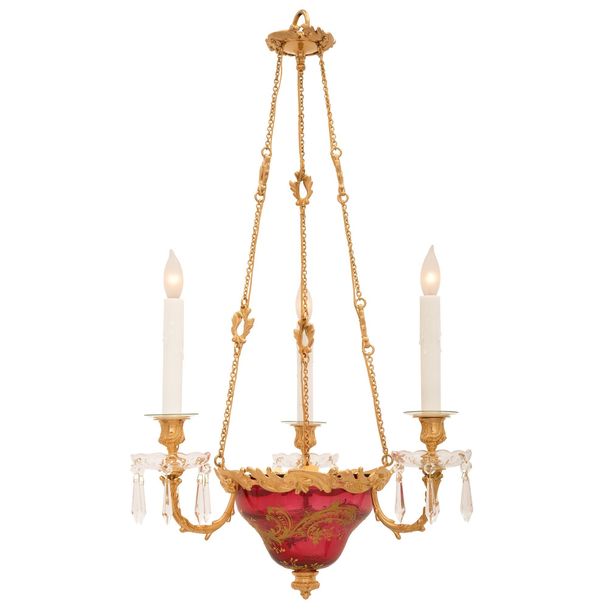 A striking French 19th century Louis XVI st. Baccarat crystal and ormolu chandelier stamped Baccarat. The three arm chandelier is centered by a charming richly chased circular mottled foliate finial below the beautifully shaped cranberry colored