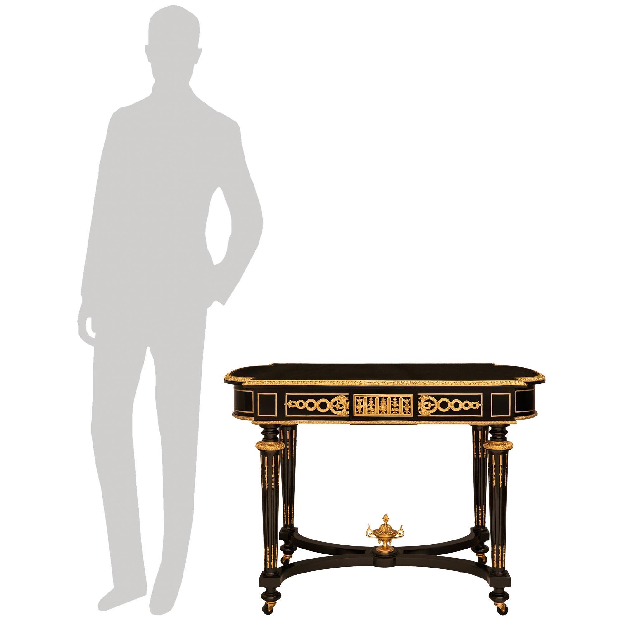 Un imponente ed elegante tavolino da centro del XIX secolo in stile Luigi XVI in ebano e Ormolu. Questo splendido tavolo si regge su quattro gambe rotonde affusolate e scanalate in ebano, dotate di rotelle e collegate da una barella a 